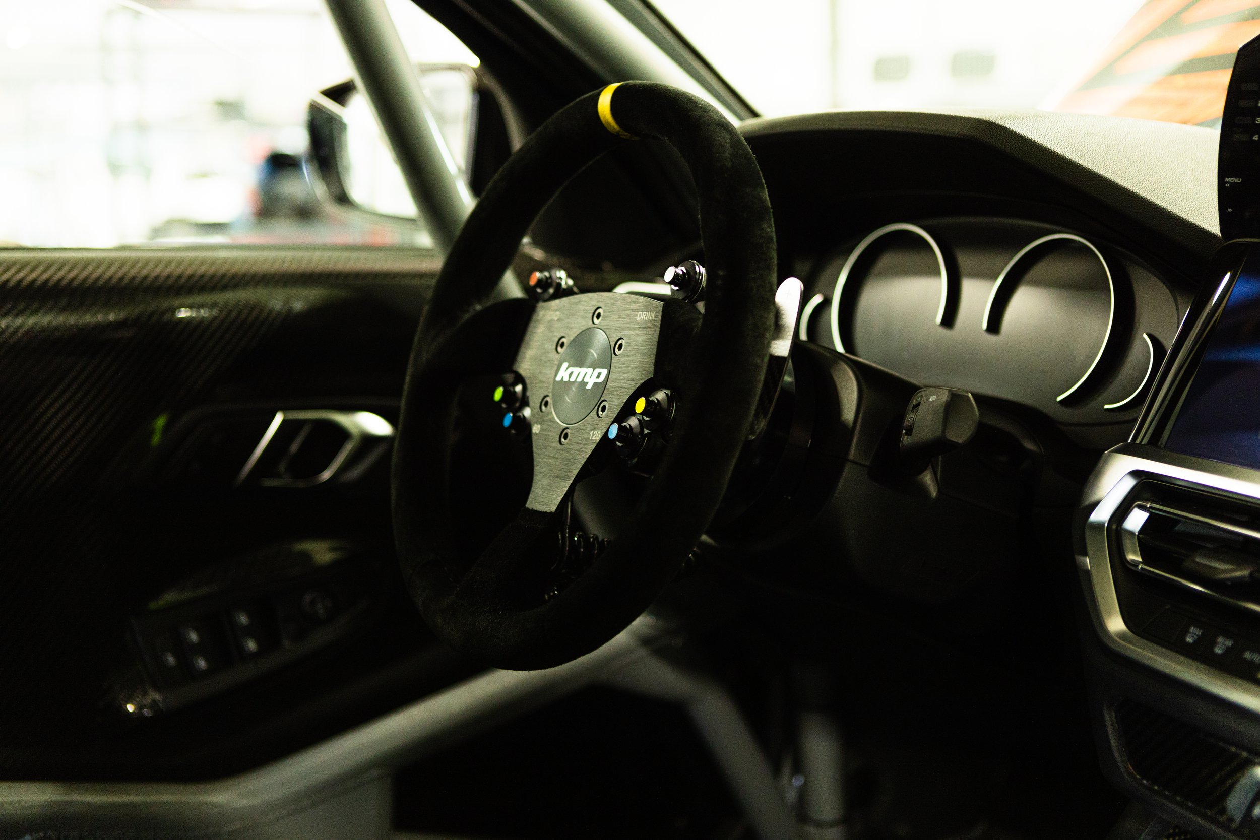 Nahaufnahme eines Lenkrads in einem Sportwagen, mit Knöpfen und einem digitalen Display im Hintergrund. Das Armaturenbrett und die Türverkleidung haben eine kohlefaserartige Oberfläche.