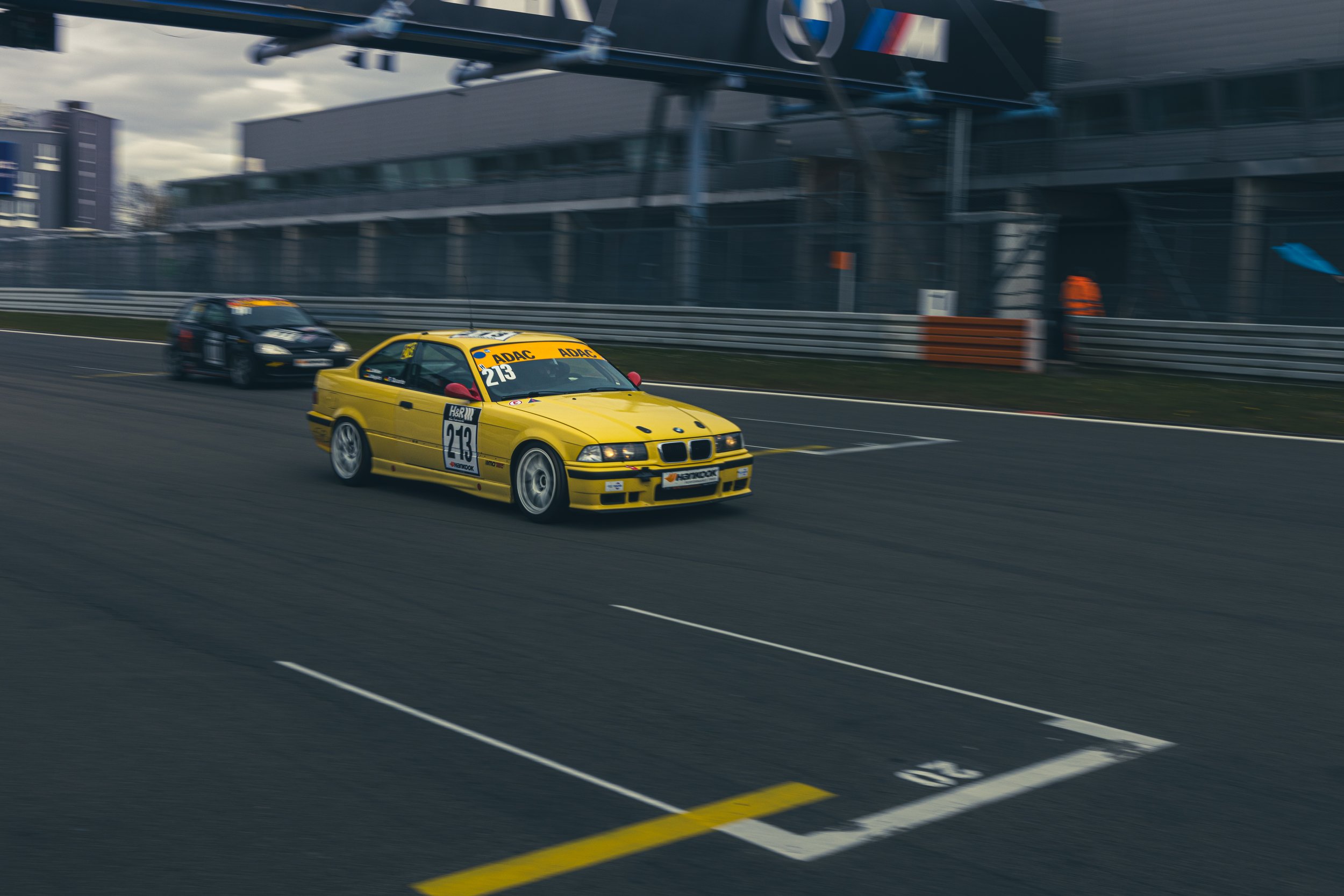 Rennwagen auf Rennstrecke, gelber BMW mit Nummer 213, ADAC Banner, zweites schwarzes Auto im Hintergrund.