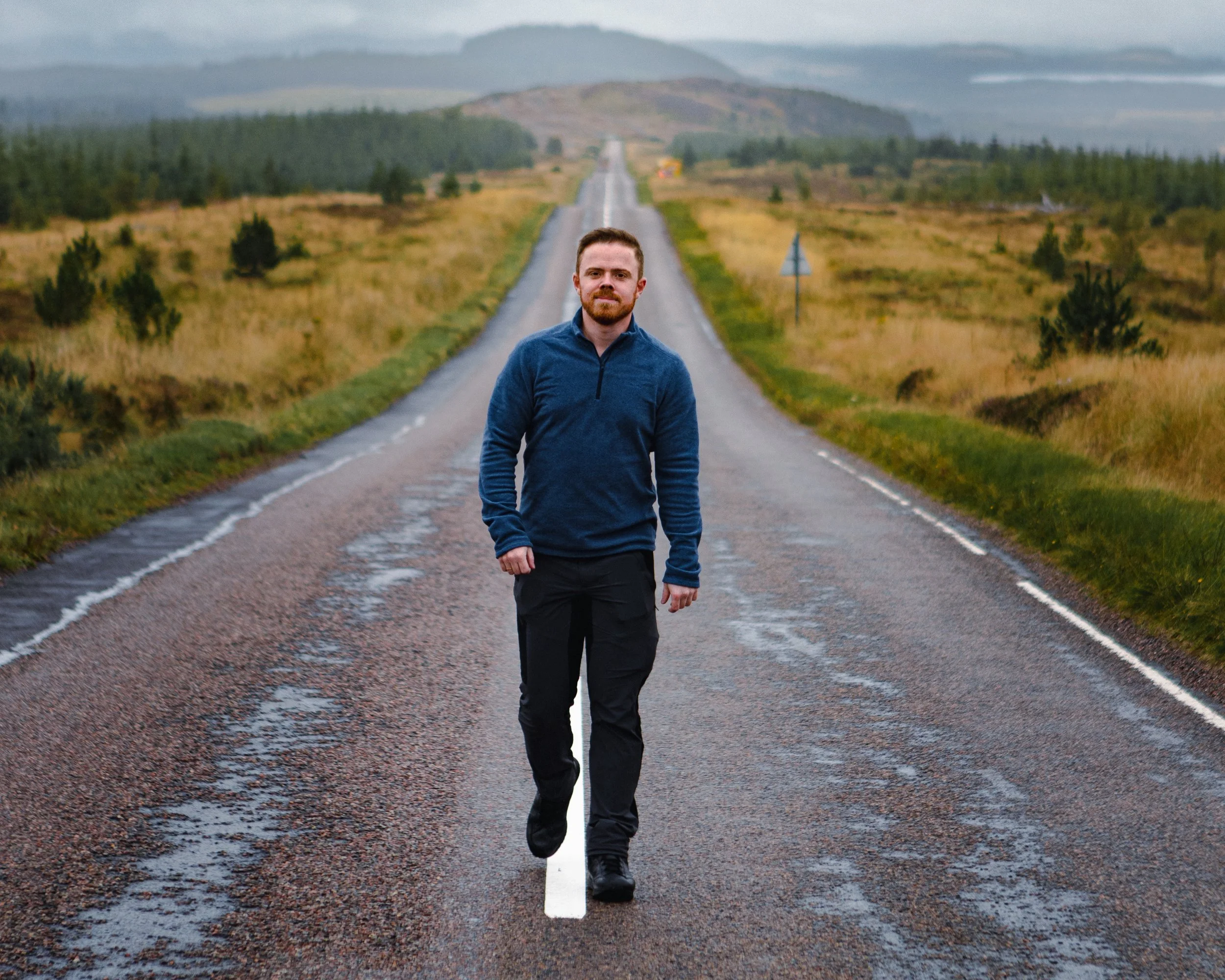 Mann in blauer Kleidung geht eine lange, gerade Straße entlang, umgeben von grünen Wiesen und Hügeln im Hintergrund.