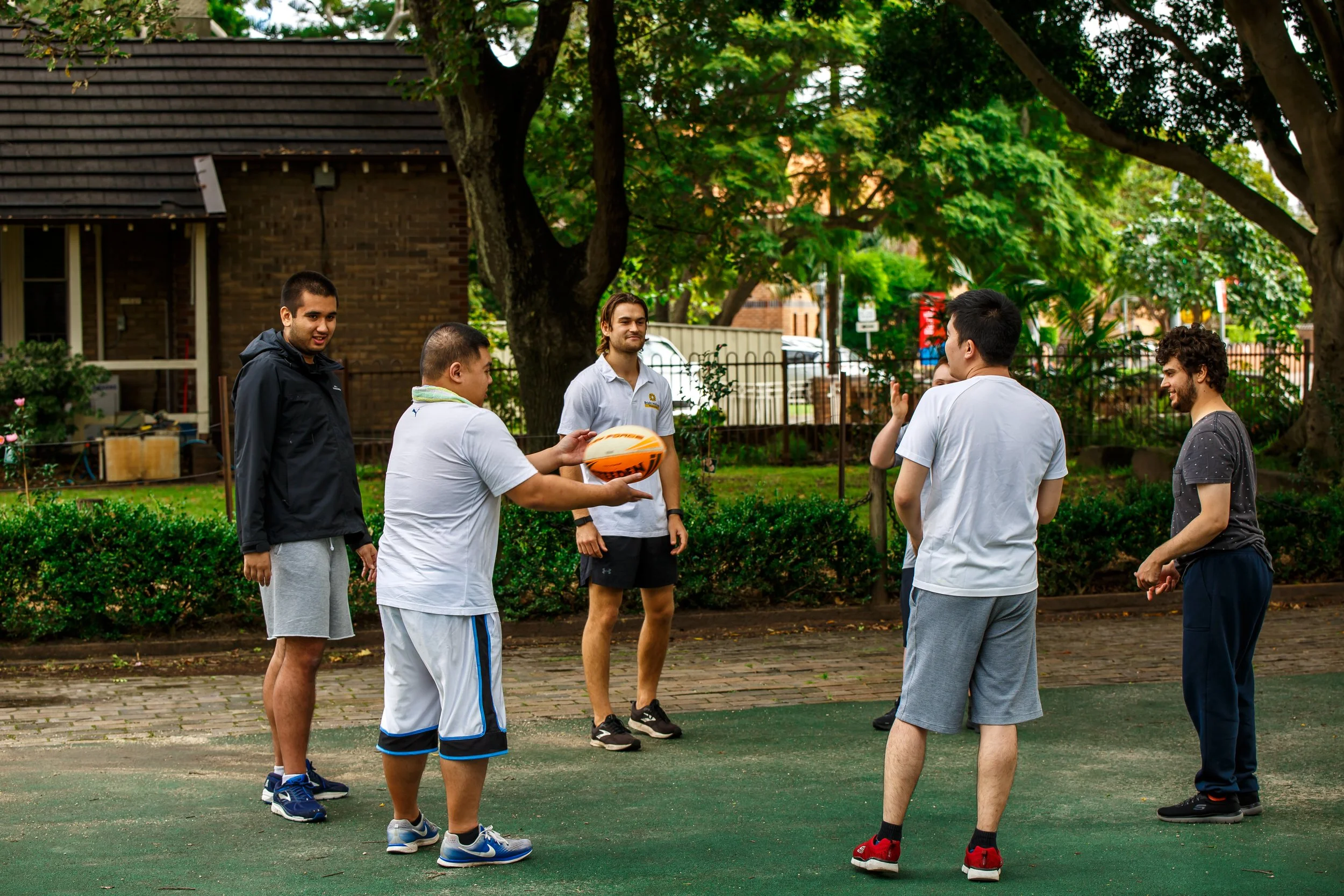 Six young men playing basketball outdoors on a court surrounded by trees and greenery.