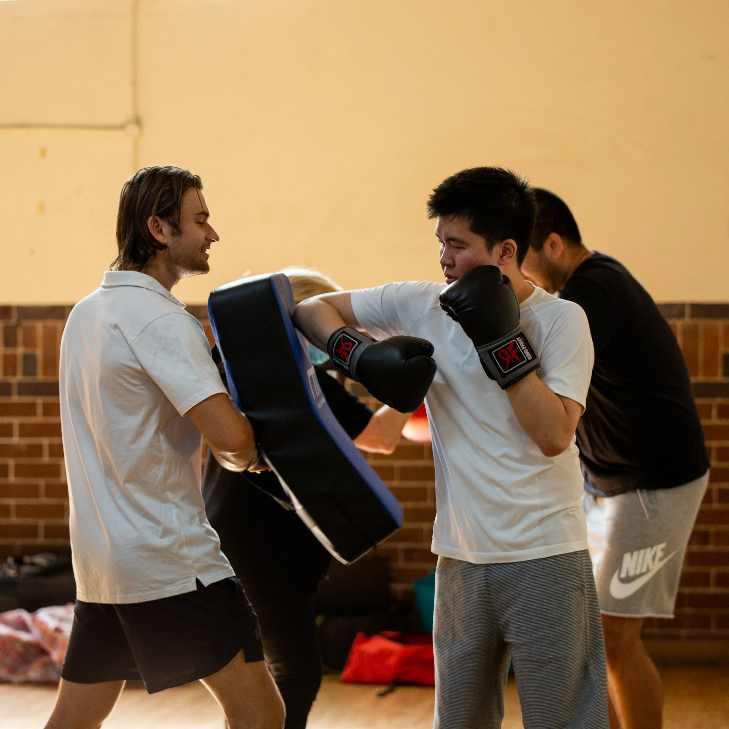 Two young men practicing boxing with one holding a punching pad while the other throws a punch, in an indoor gym with brick walls.