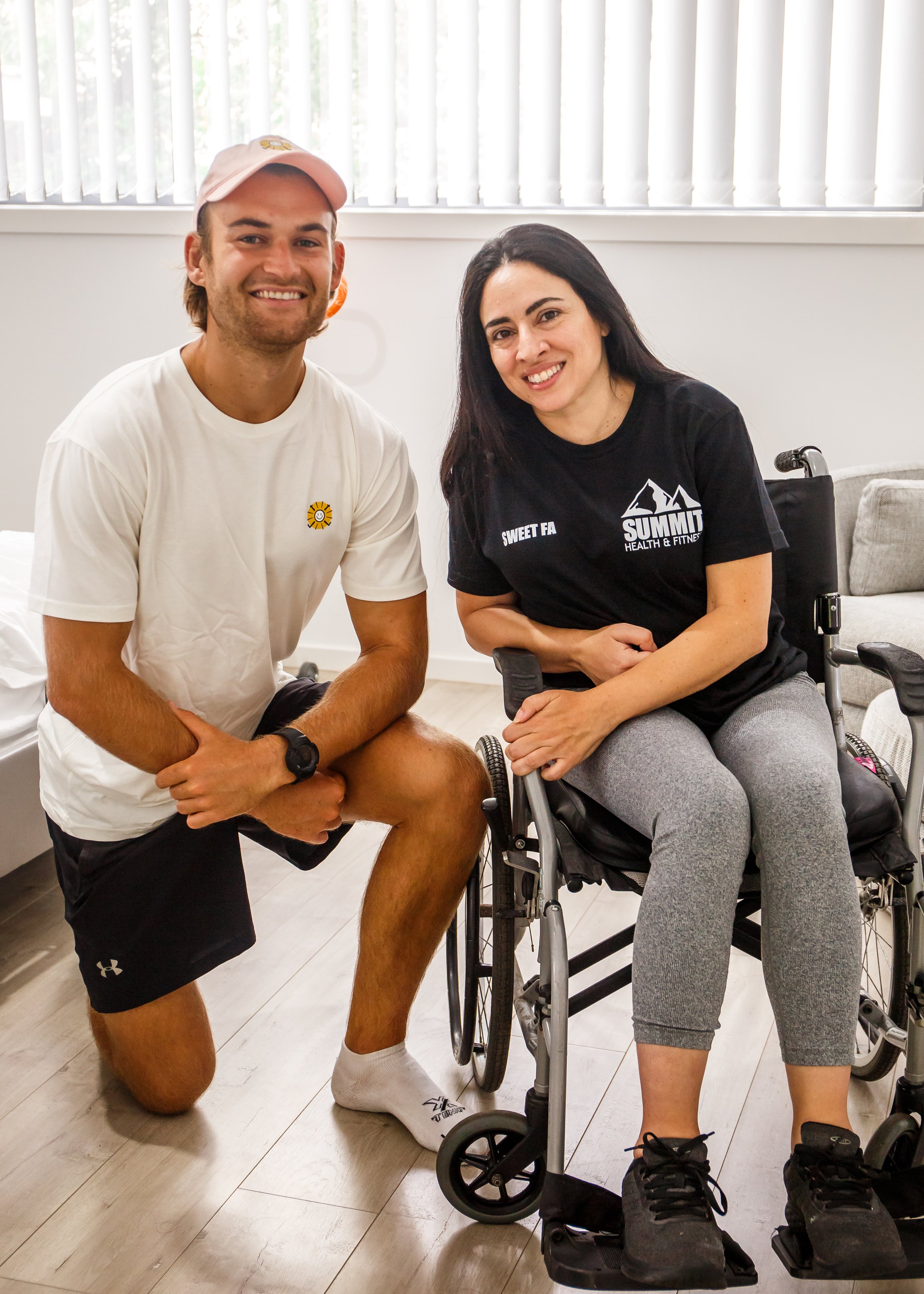 A smiling man kneeling next to a woman in a wheelchair in a room with white walls and vertical blinds on the window, both looking at the camera.