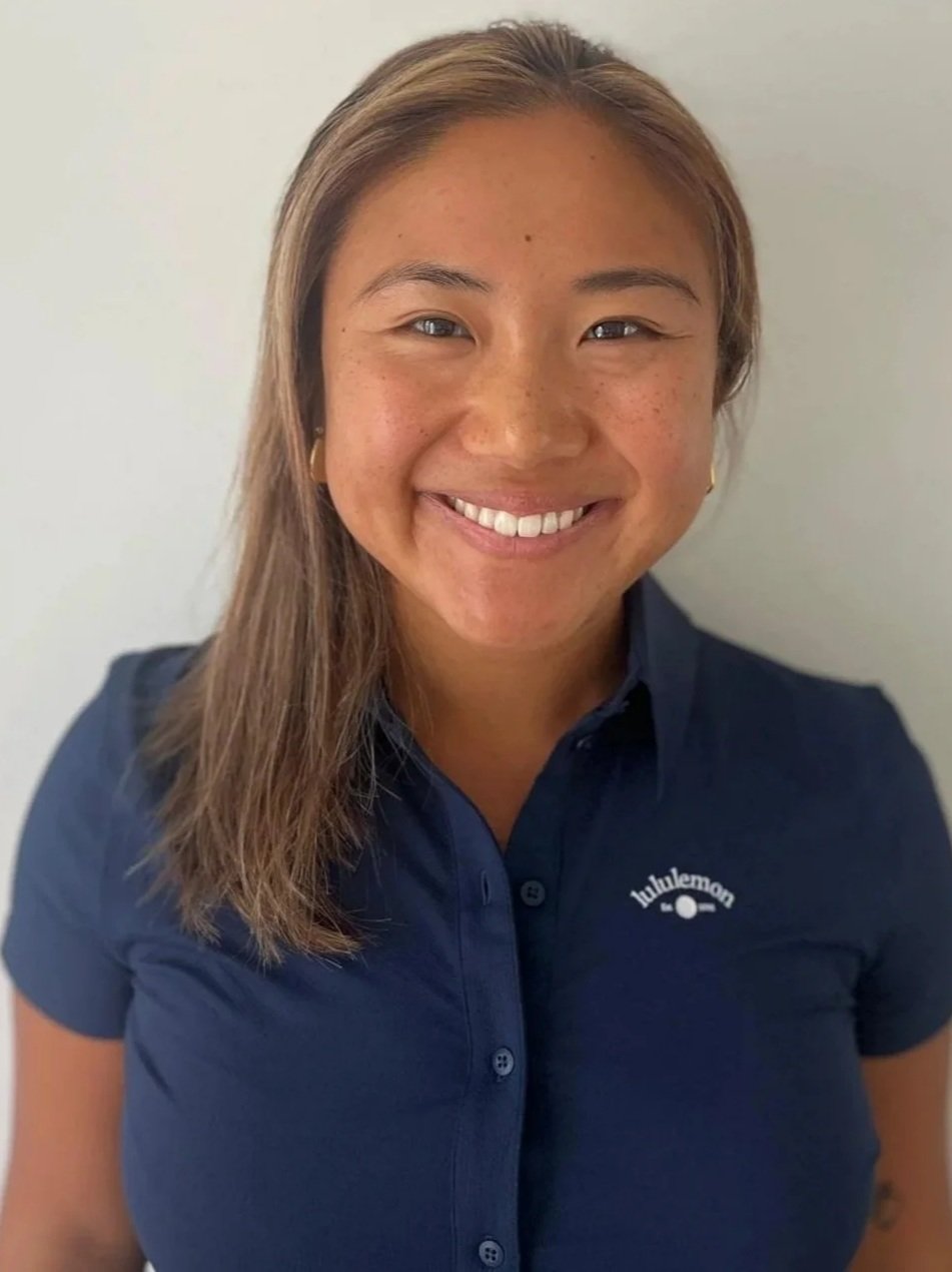 A young woman with long reddish-brown hair and blue eyes smiling at the camera, wearing a white shirt and a delicate necklace, against a light blue wall background.