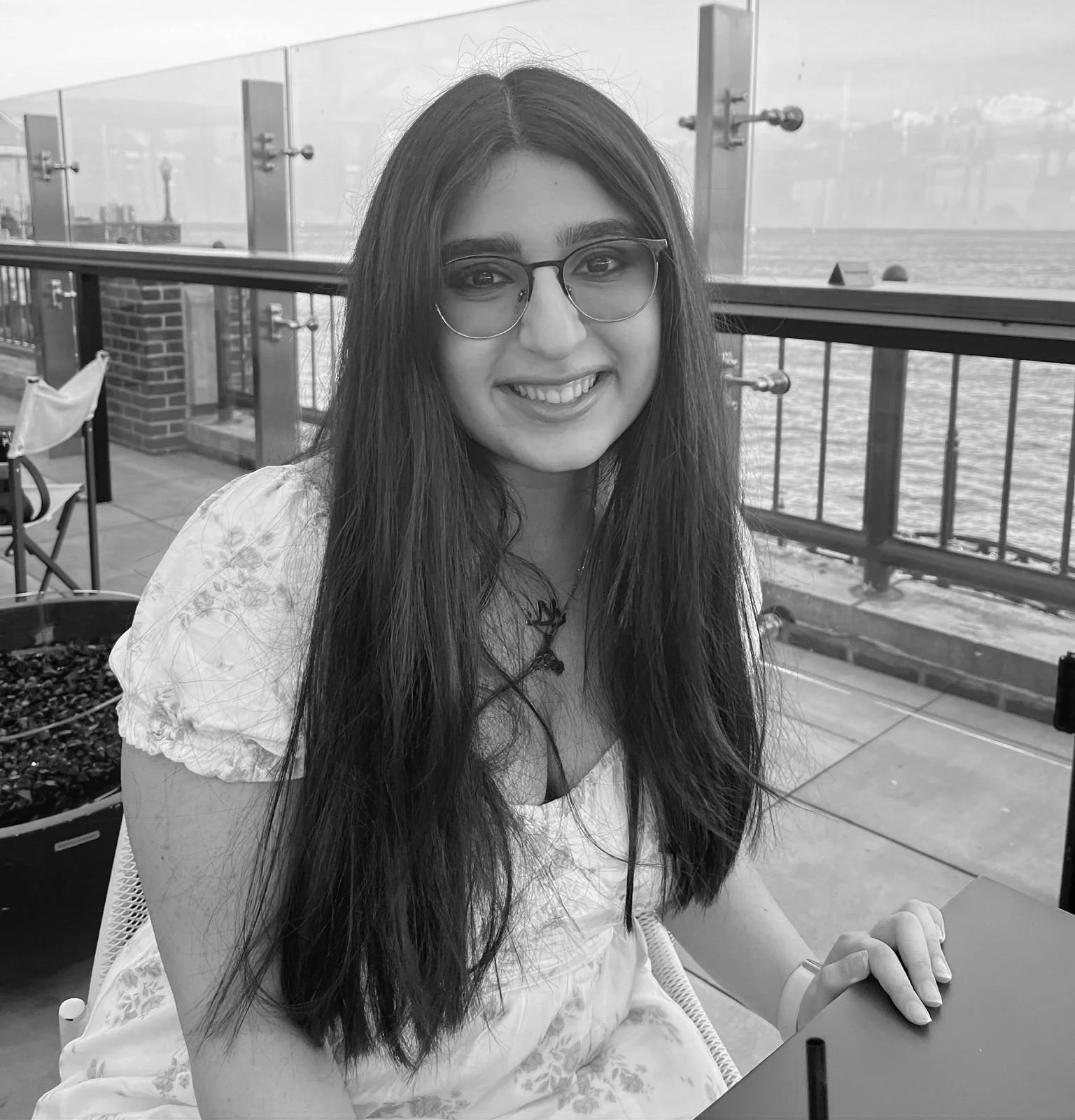 A young woman with long dark hair and glasses sitting at an outdoor table near a body of water, smiling at the camera.