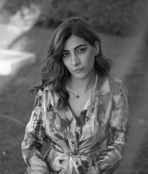 A woman with wavy hair and a serious expression, wearing a patterned blouse, outdoors with trees and grass in the background.
