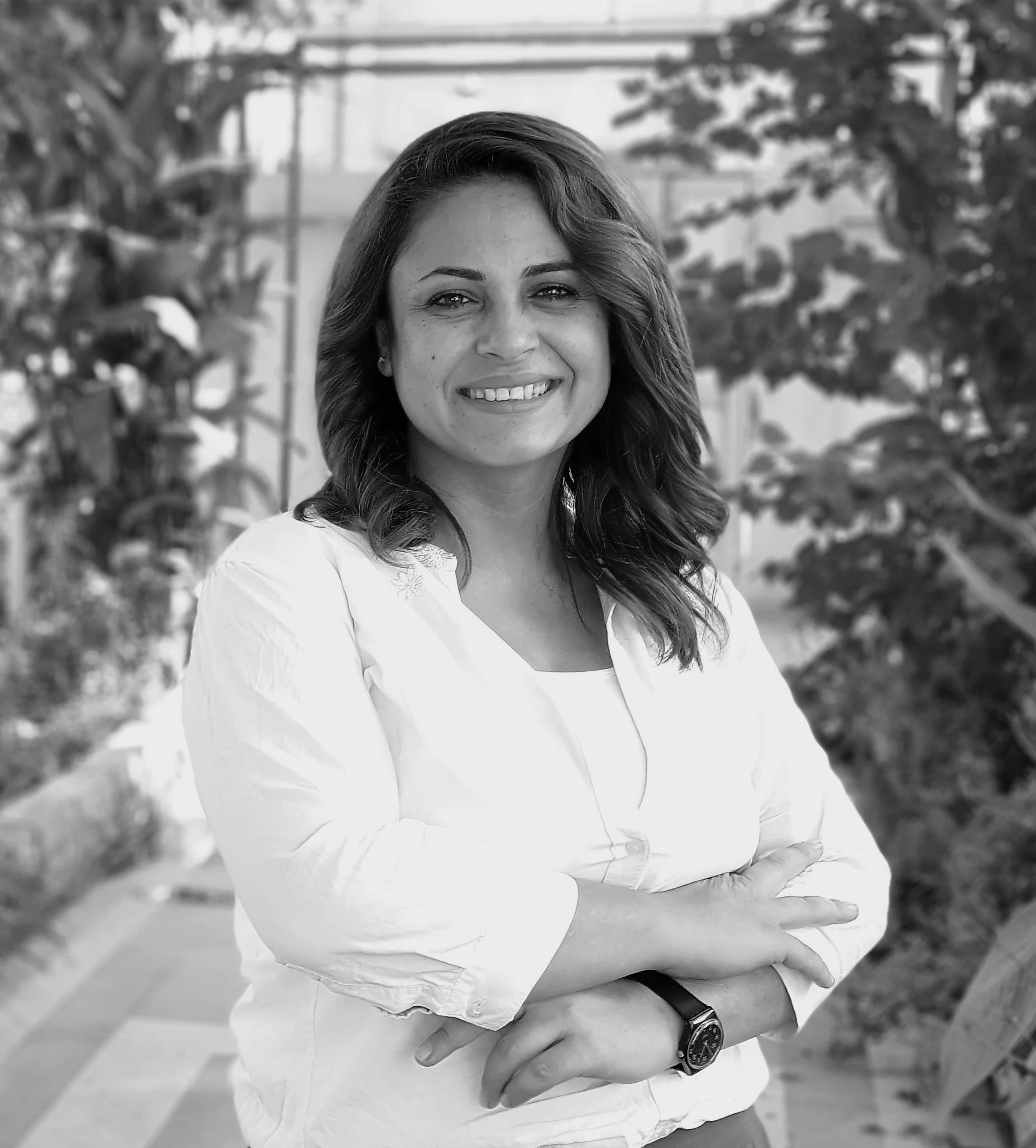 A woman with shoulder-length hair smiling, standing outdoors with trees and a bridge in the background, wearing a light-colored blouse and a watch on her left wrist.
