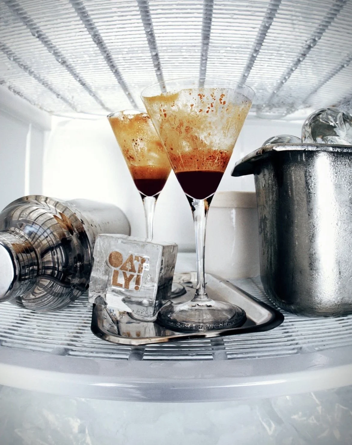 Two glasses with brown and orange layered drinks inside a refrigerator shelf, along with a metallic shaker, a block of ice with a logo, and some ice cubes in a metal container.