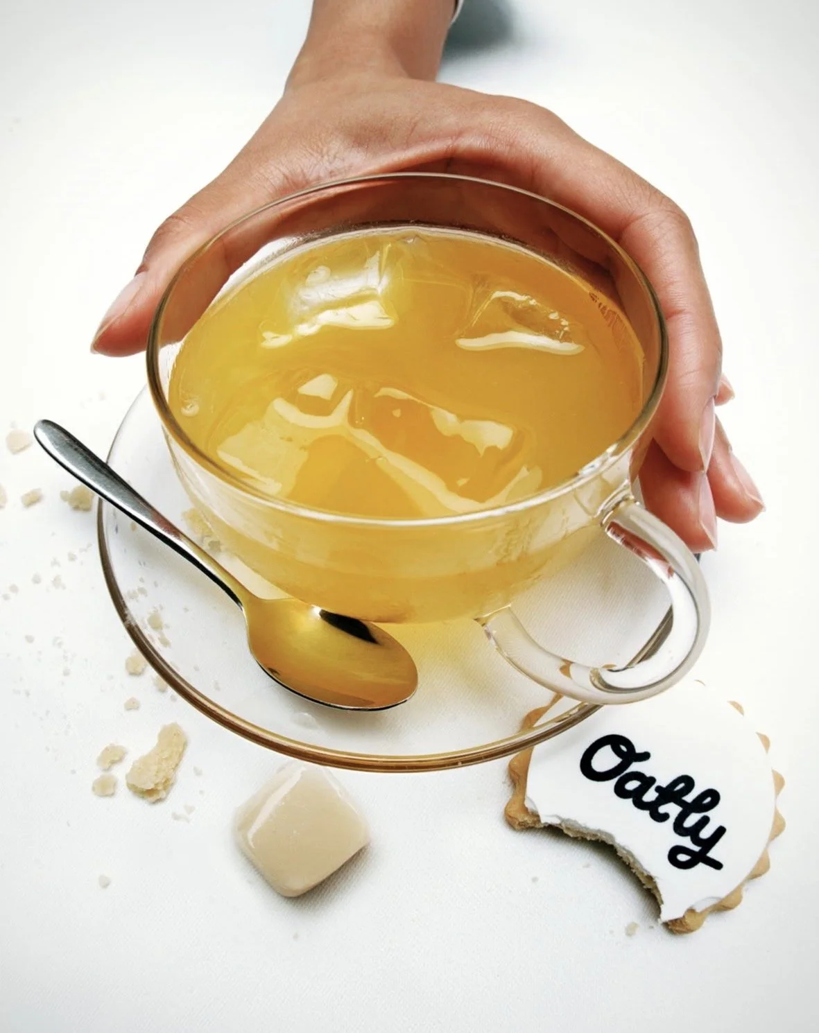 A hand holding a glass cup filled with iced tea on a white surface. There is a small metal spoon on a saucer, some broken cookies, and a cookie with white icing and the word 'Oatly' written on it.
