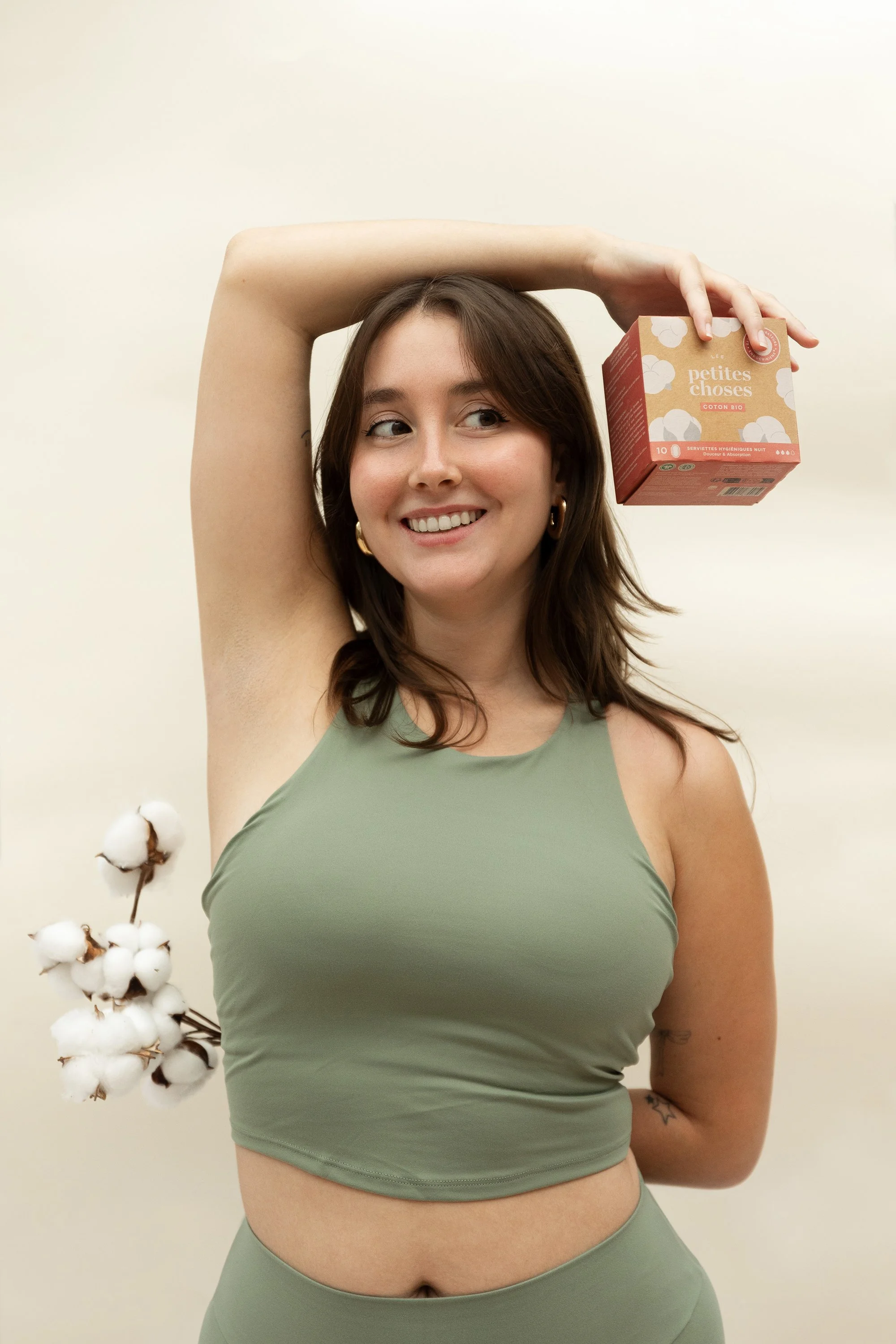 Une jeune femme souriante en tenue de sport vert, tenant un petit carton de serviettes hygiéniques bio dans la main droite au-dessus de sa tête, avec une branche de coton dans le dos.