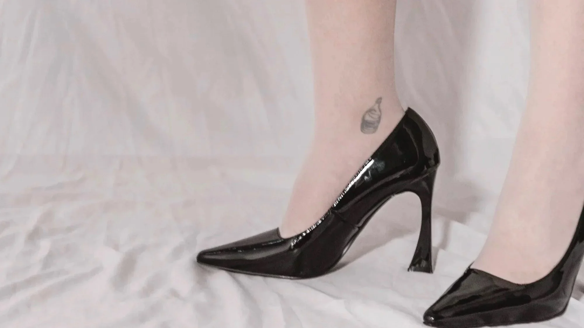 Pied de femme portant des talons aiguilles noirs en plastique brillant, avec tatouage de bouteille d'eau sur la cheville gauche, sur un drap blanc.