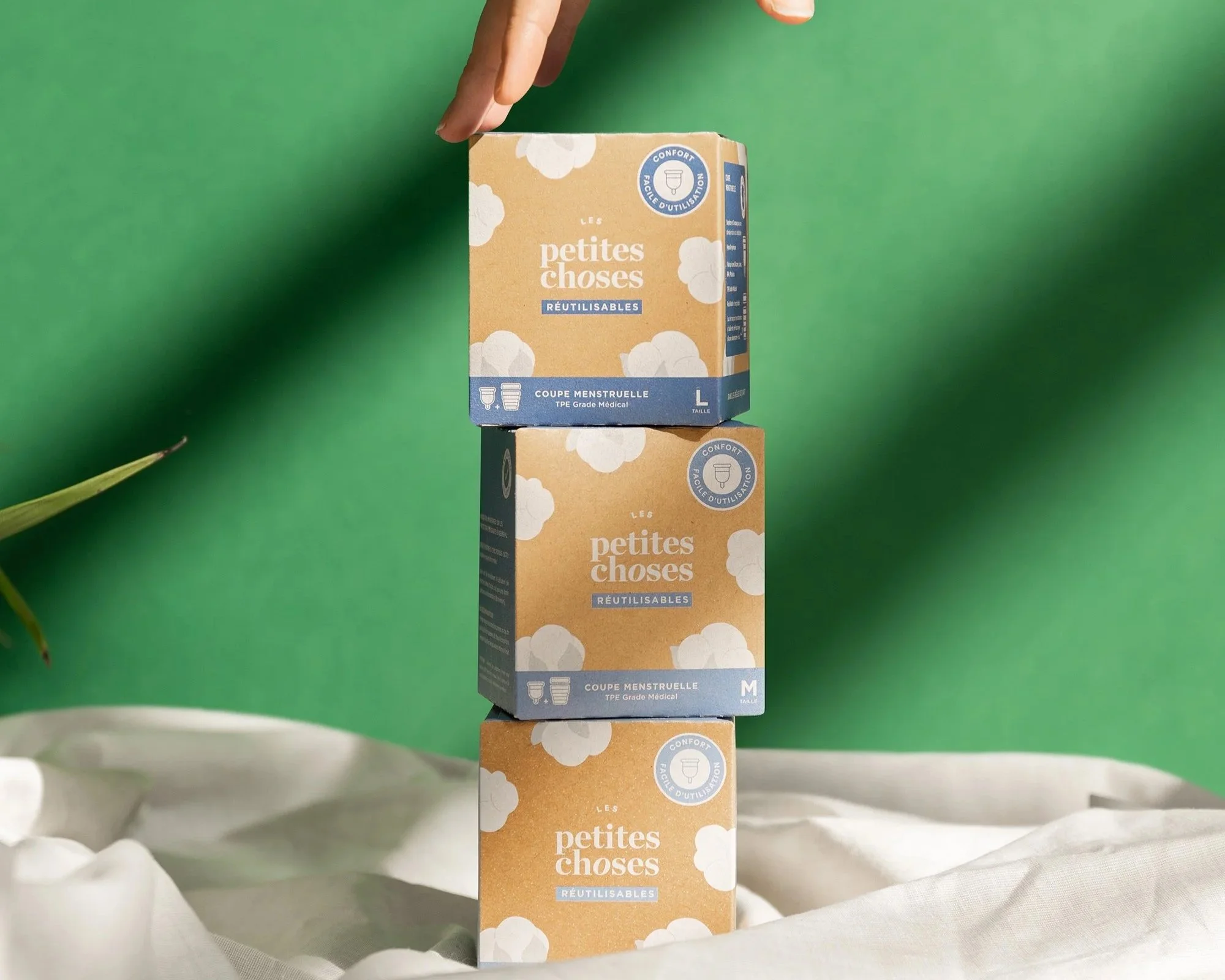 Trois boîtes de tampons réutilisables pour règles empilées verticalement, avec une main les soutenant, contre un fond vert avec un drapé blanc en dessous.
