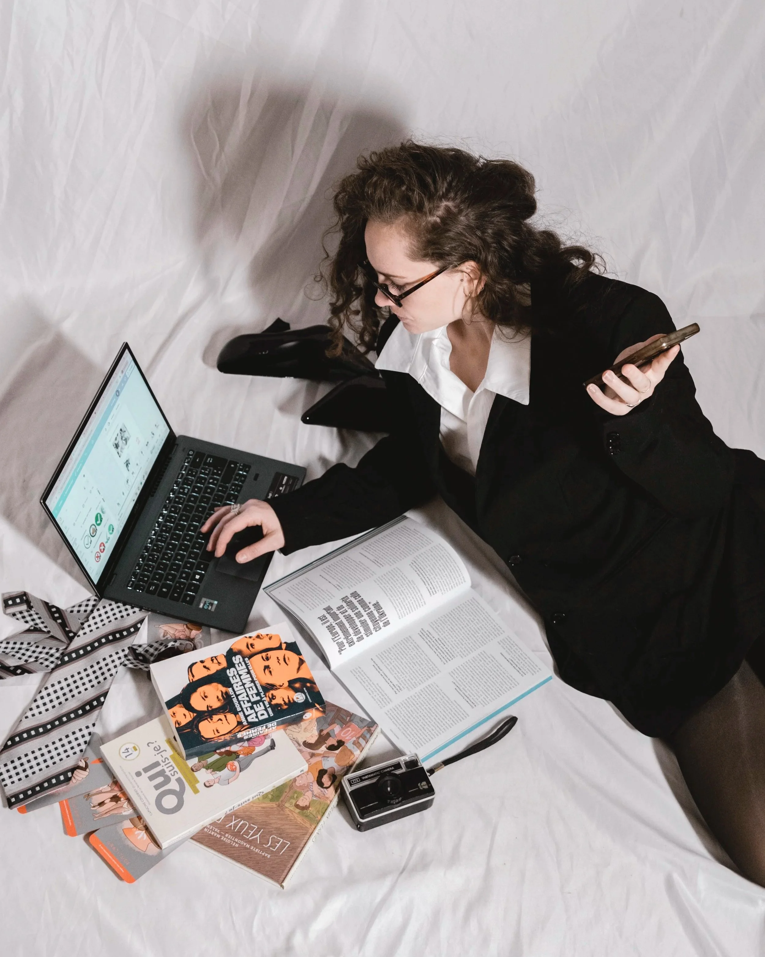 Femme allongée sur un lit, utilisant un ordinateur portable, entourée de livres, d’un appareil photo, d’un téléphone et d’une carte mémoire.