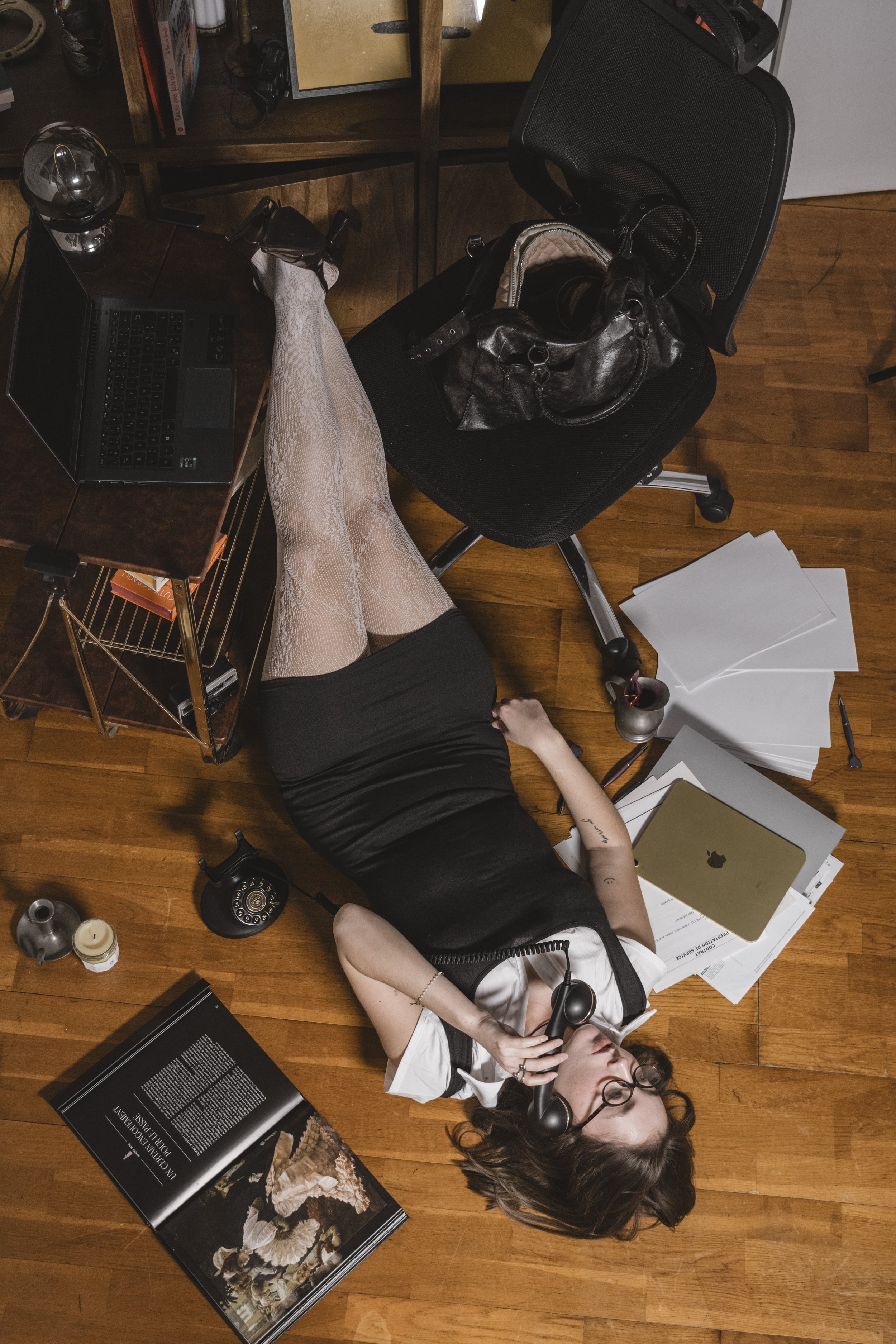 Une femme allongée sur un sol en bois, parlant au téléphone, entourée de documents, d'un ordinateur portable, et de plusieurs objets de bureau. Elle porte des lunettes, une robe noire et blanche, et des collants en dentelle.