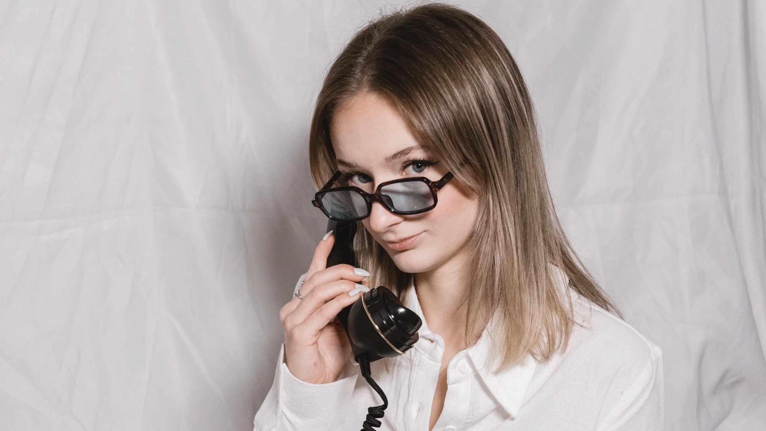 Une femme avec des lunettes de soleil, tenant un téléphone rétro, portant une chemise blanche, se regarde avec un léger sourire.