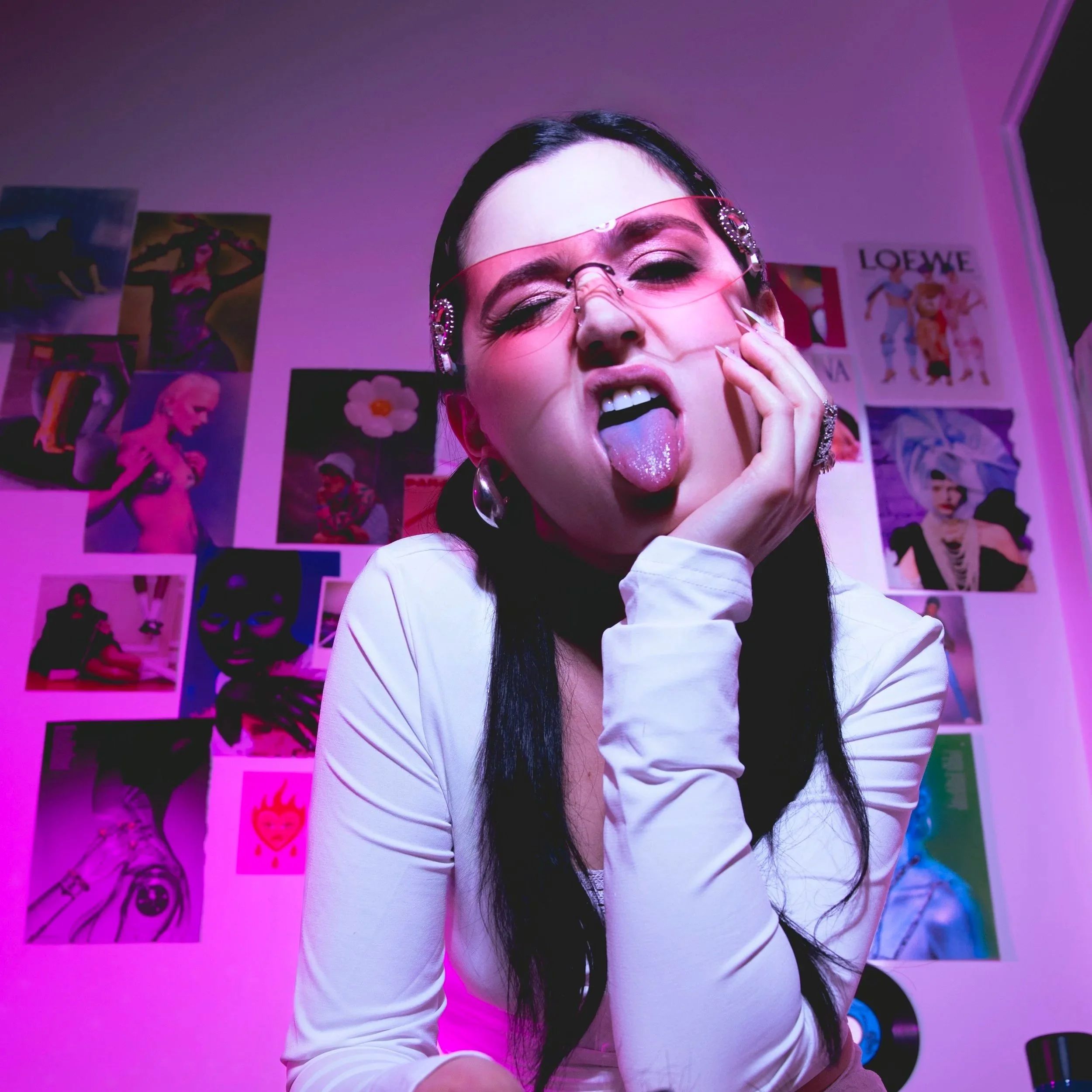 Jeune femme avec lunettes roses et boucle d'oreilles, faisant un geste avec sa langue et sa main dans une pièce décorée avec des posters et des affiches colorés.