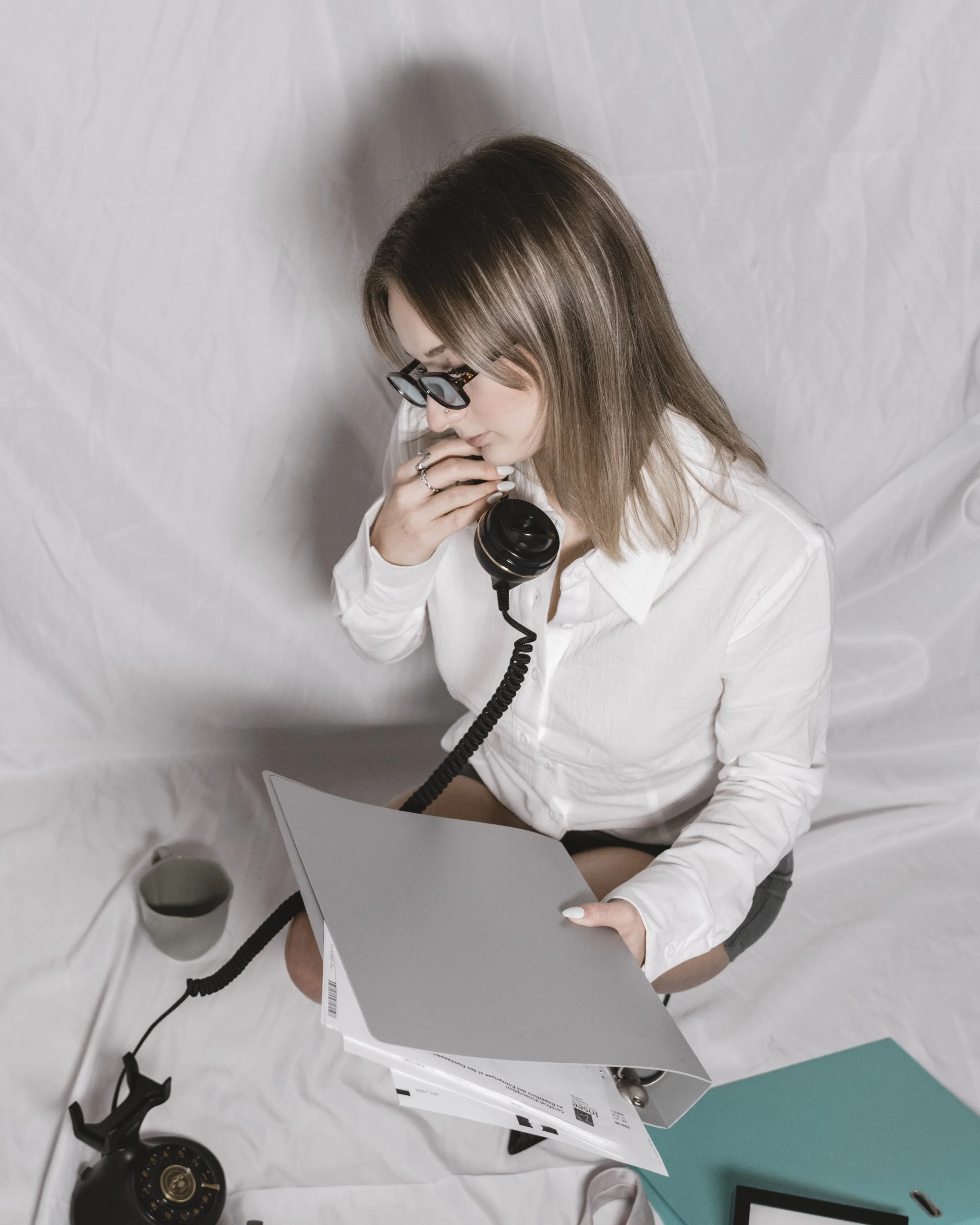 Une femme avec des lunettes assise sur un fond blanc, tenant un téléphone avec un récepteur branché, avec des documents et un mug autour d'elle.