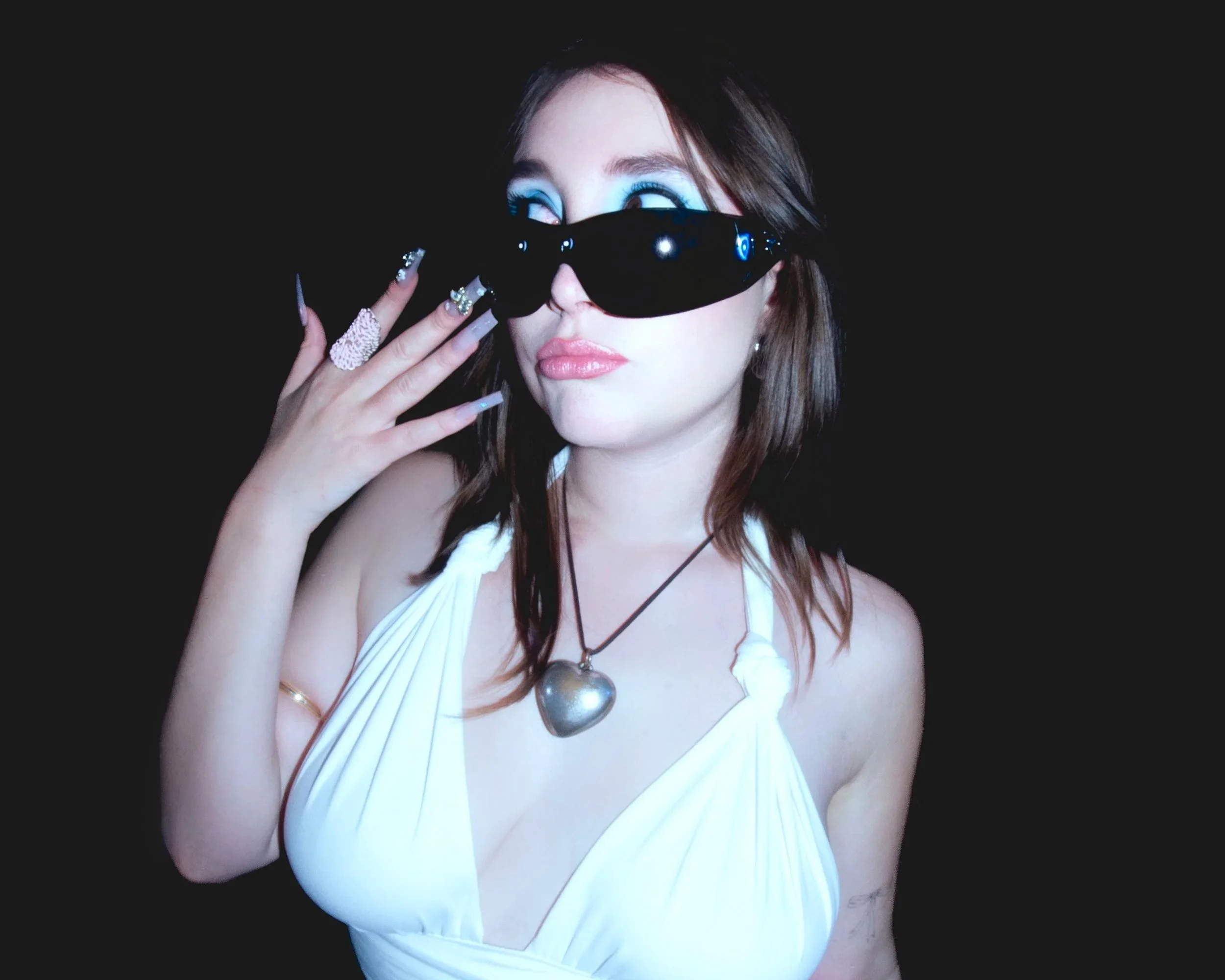 Jeune femme portant des lunettes de soleil noires, une robe blanche à baillettes avec des épaules dénudées, un collier en forme de cœur en métal argenté, et une manucure élaborée, pose contre un fond noir.