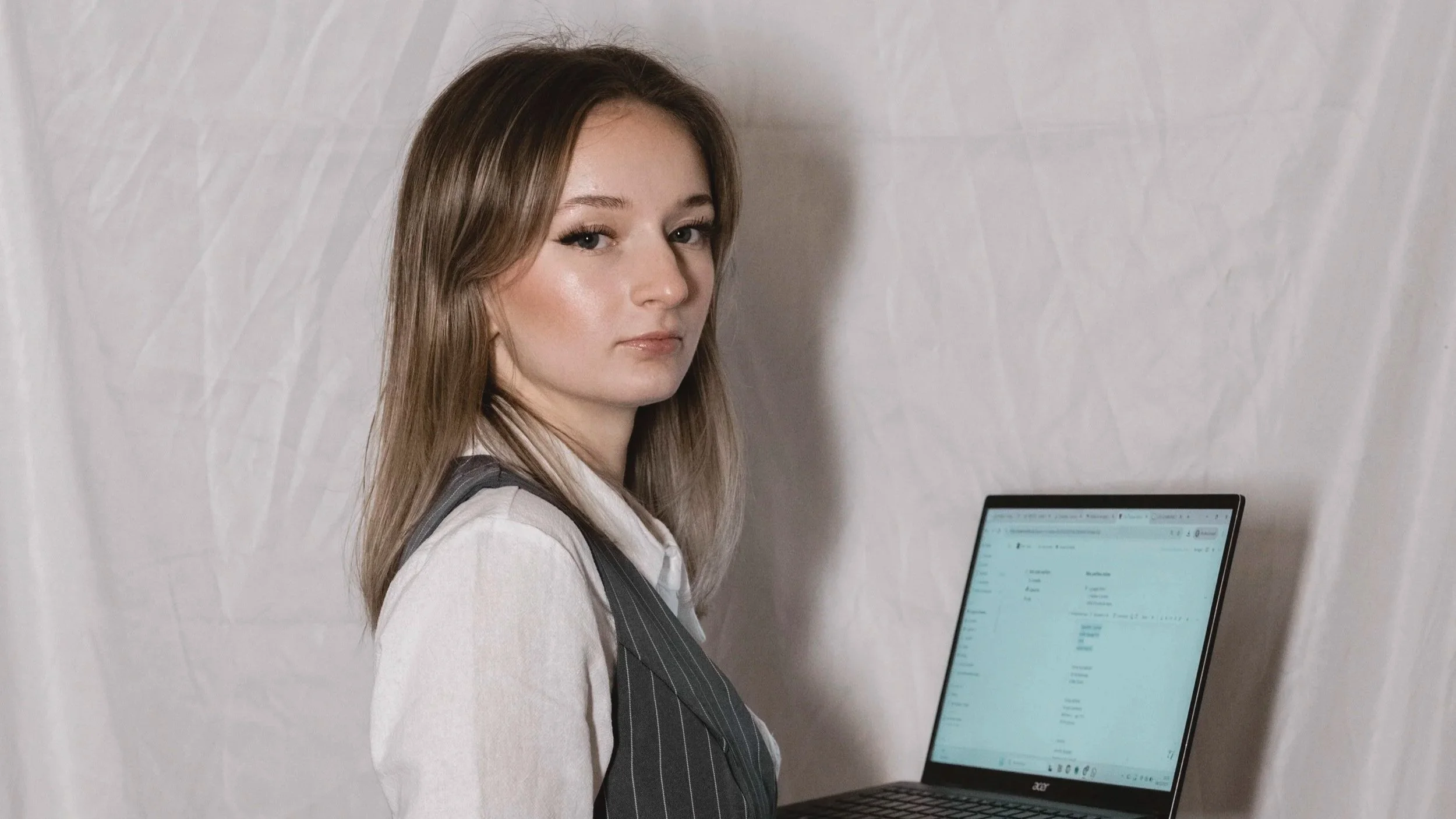 Une femme avec des cheveux bruns, portant une chemise blanche et un gilet gris à rayures, se tenant à côté d'un ordinateur portable et regardant l'objectif, contre un fond blanc.