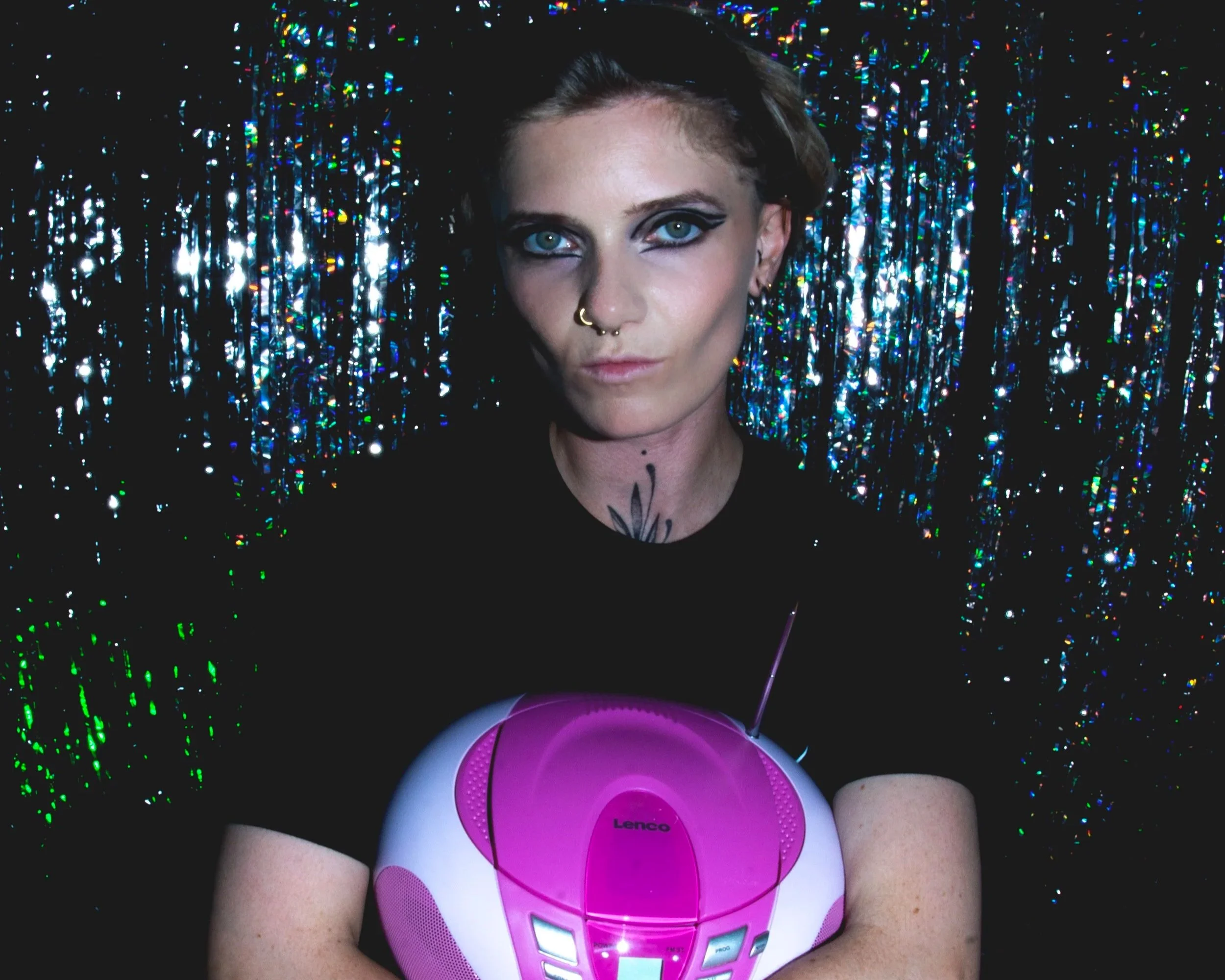 Une femme avec un regard intense, des yeux bleus, des yeux soulignés de maquillage noir, porte un piercing au nez, tient un radio-cassette rose, avec un fond de rubans holographiques brillants.
