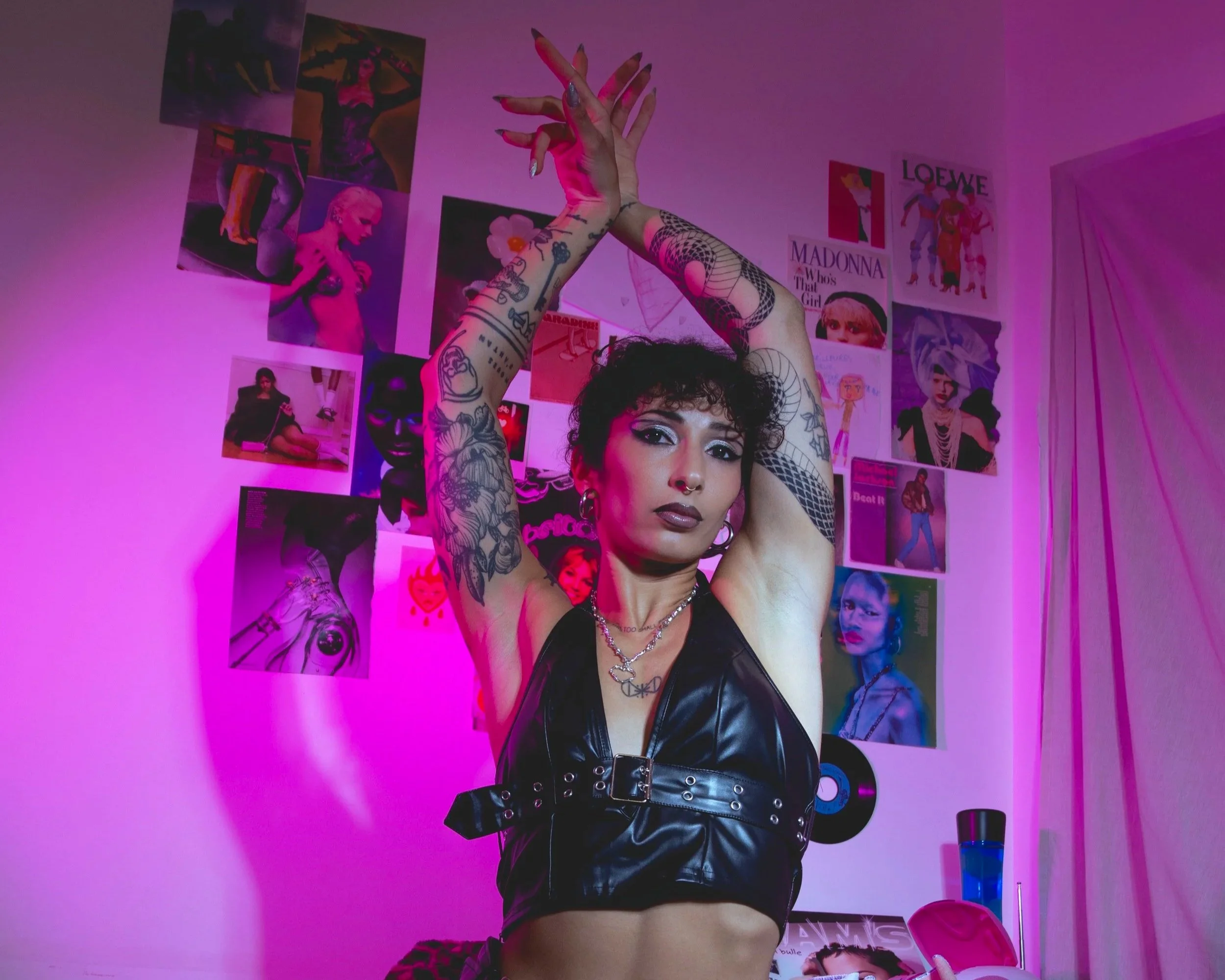 Une femme avec tatouages, portant une veste en cuir noire, pose avec les bras levés dans une pièce décorée avec des posters de célébrités et des images colorées.