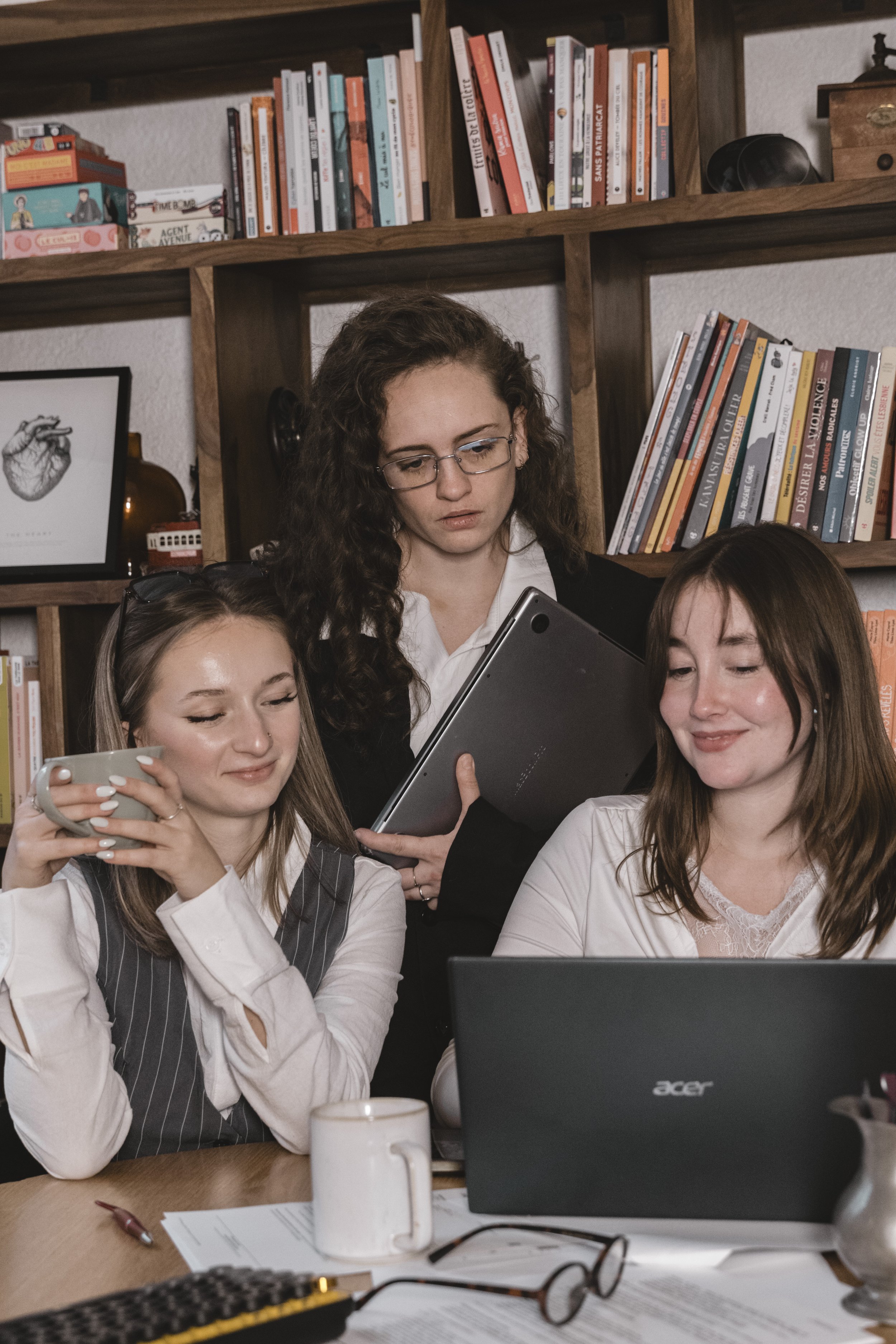 Trois jeunes femmes autour d'une table, travaillant ensemble avec des ordinateurs portables et des livres, dans une bibliothèque ou un bureau, avec des étagères remplies de livres en arrière-plan.