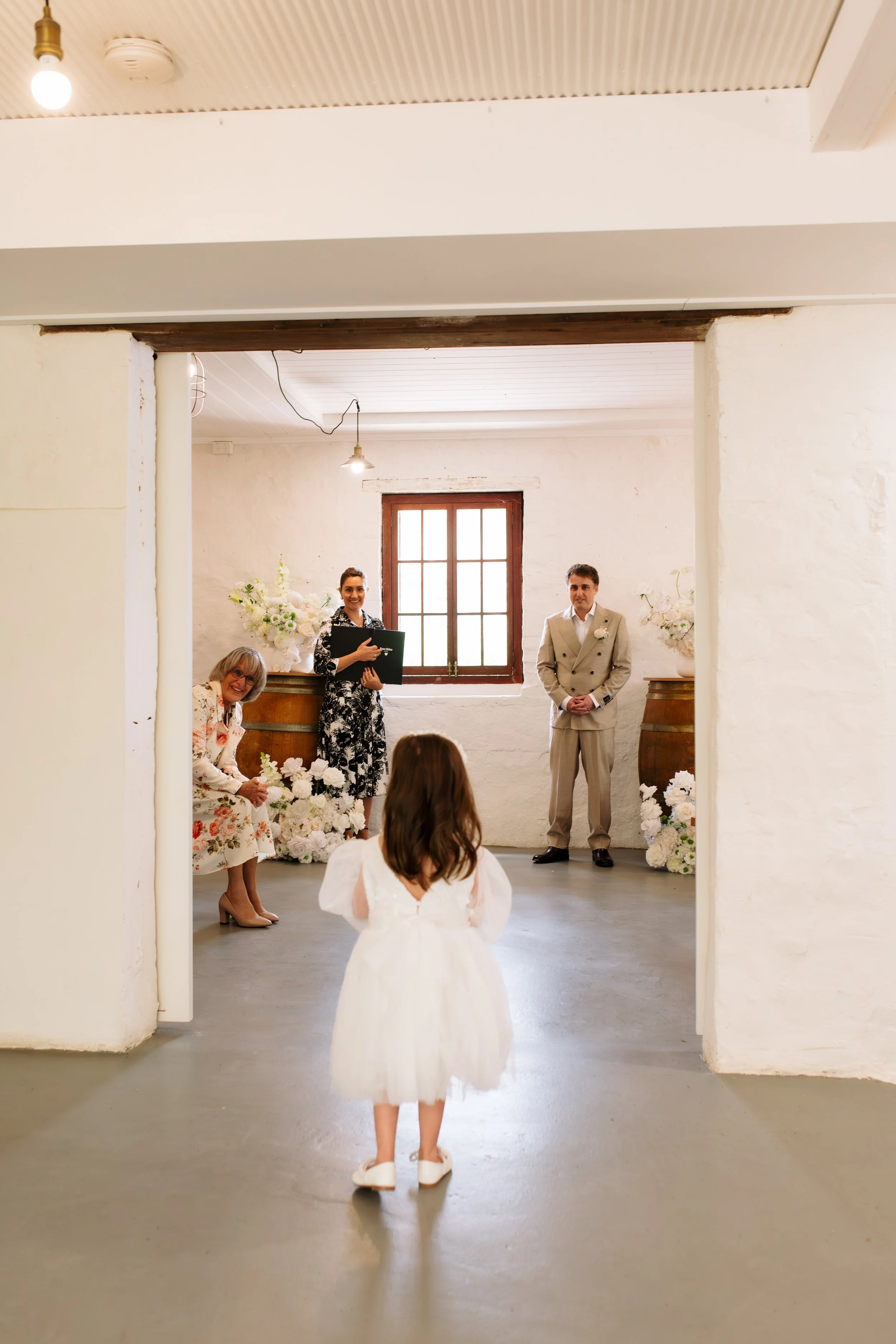 A bride in a white dress is facing three people standing in a rustic room with white walls and wooden accents, during a wedding ceremony.