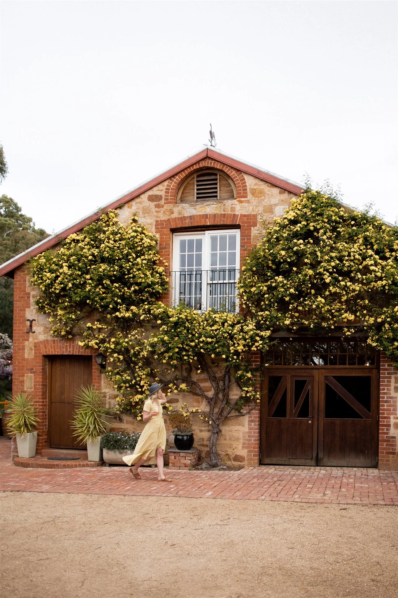 A woman in a yellow dress and wide-brimmed hat walking past a rustic two-story house with a stone and brick facade, featuring yellow flowering plants climbing the front, a wooden door, and a garage with sliding doors.
