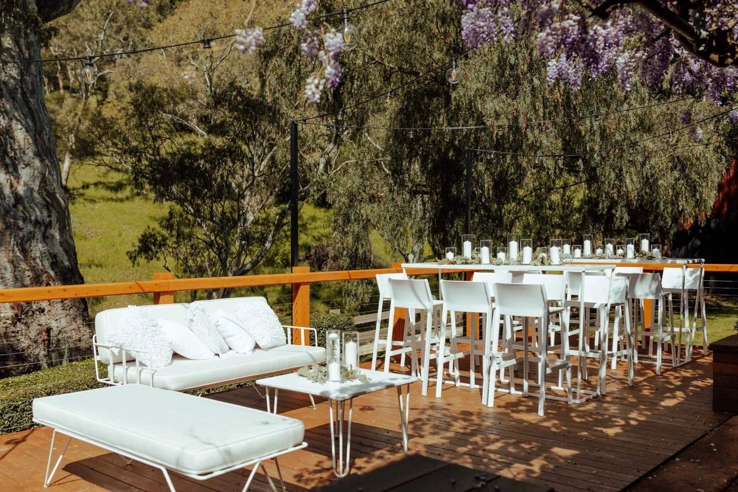 Outdoor deck decorated for a gathering, with white furniture, a large dining table with candles, a sofa and ottoman, and string lights above, surrounded by trees and nature.