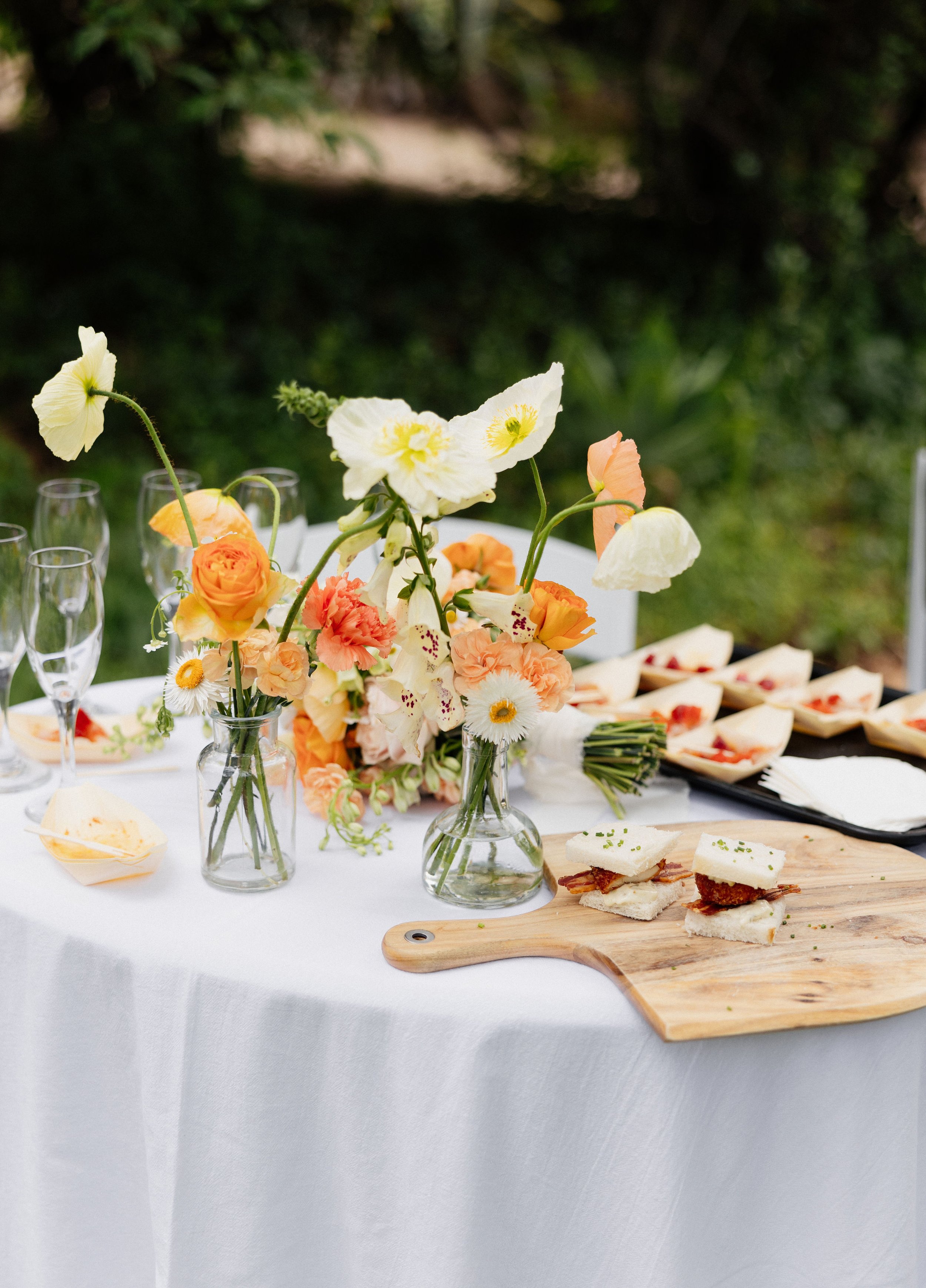 A table with a white tablecloth, decorated with a bouquet of peach, yellow, and white flowers in glass vases, and set with glasses, plates, and snacks including sandwiches and hors d'oeuvres, outdoors with greenery in the background.