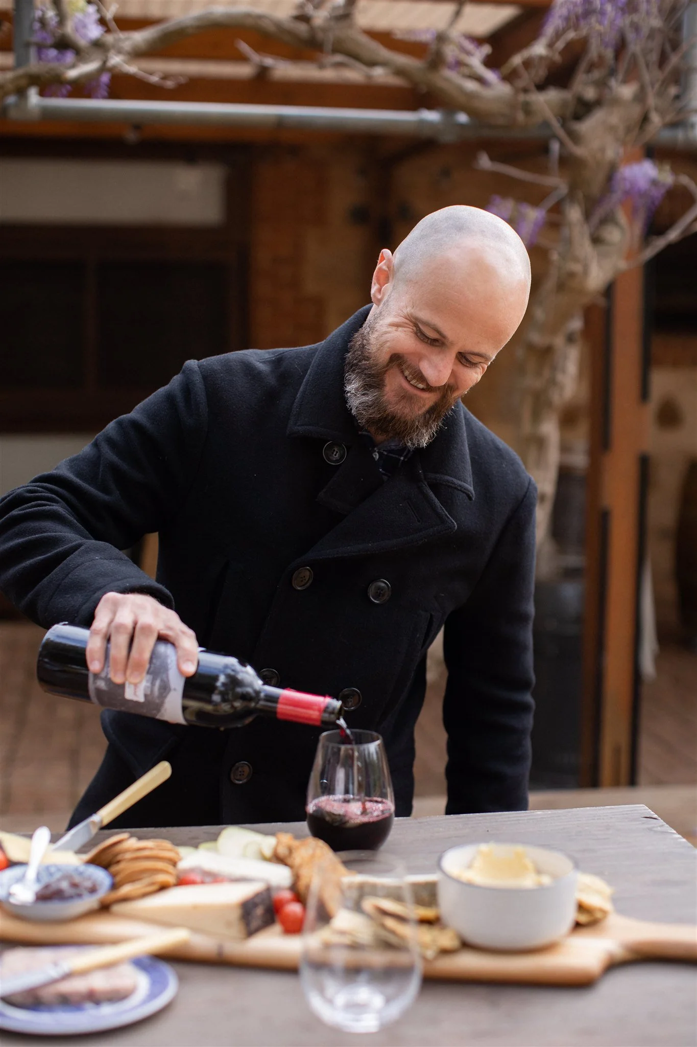 A man with a beard and a shaved head pouring red wine into a glass on a table with cheese, crackers, and fruit, indoors with a wooden background and a tree with purple flowers.