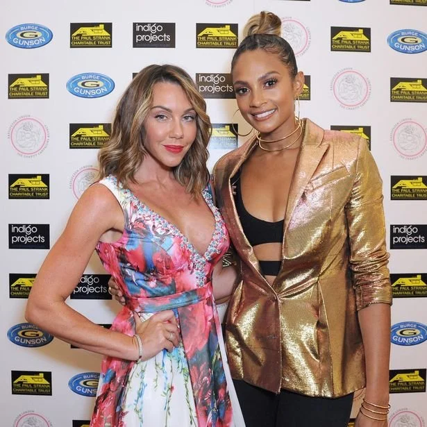 Two women are standing together in front of a step-and-repeat backdrop. One woman is wearing a colorful floral dress, and the other is wearing a metallic gold blazer over a black top. They are smiling at the camera.