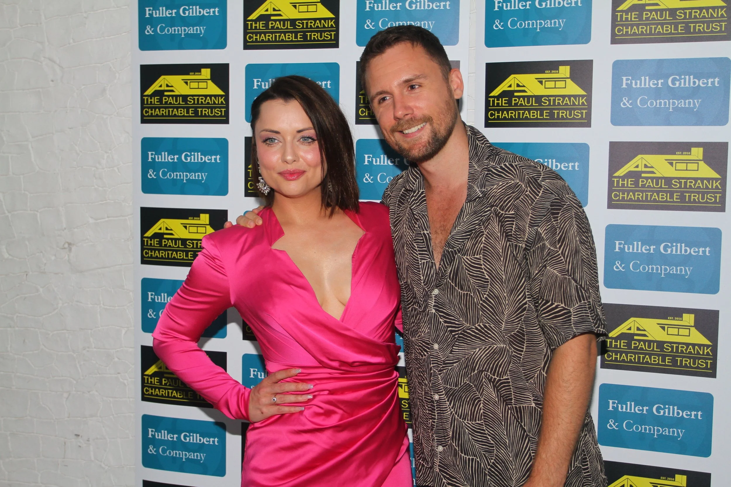 A woman in a bright pink satin dress and a man in a patterned shirt posing together at an event with a background displaying logos for 'Fuller Gilbert & Company' and 'The Paul Strank Charitable Trust'.