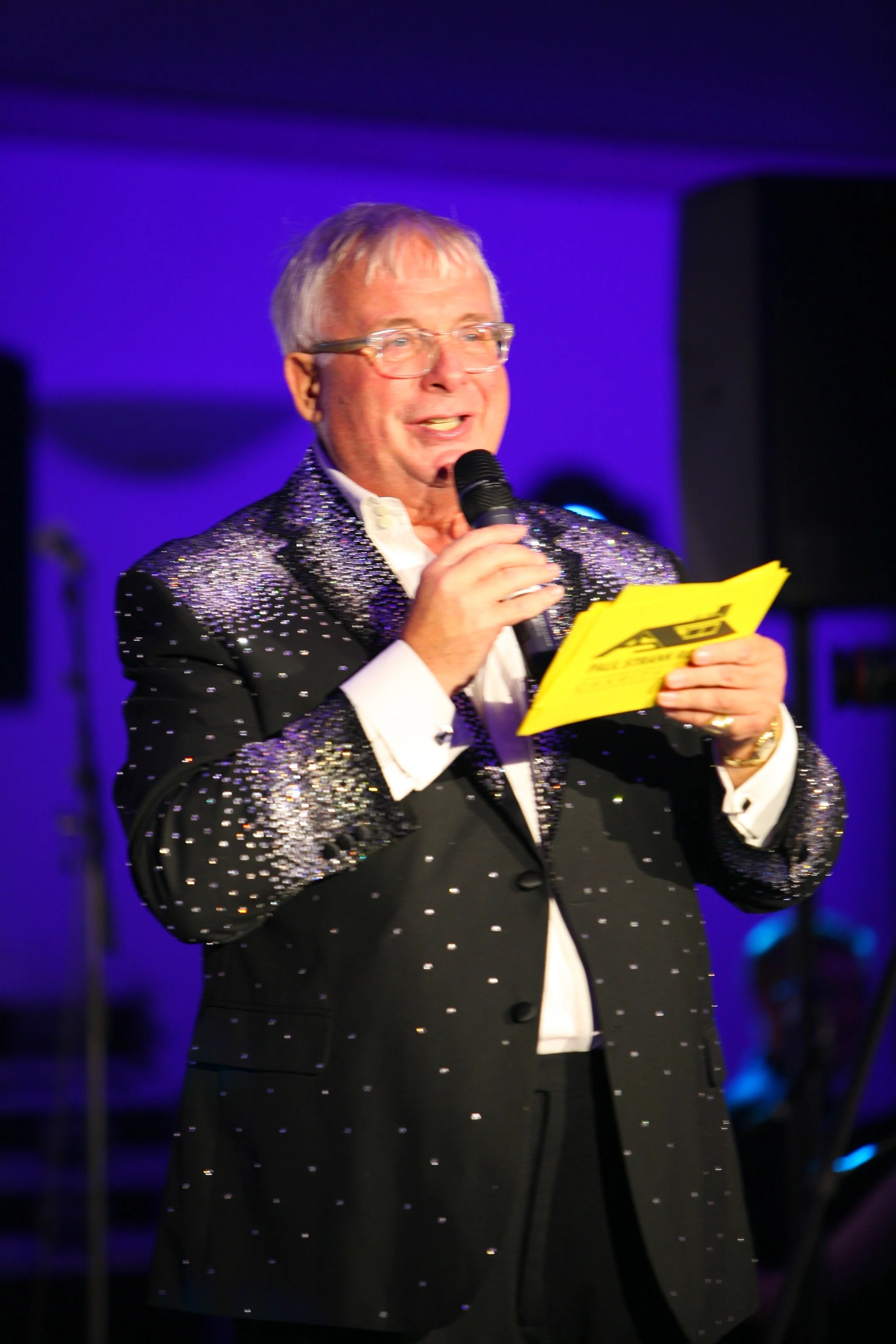 A man in a black blazer glittering with rhinestones is speaking into a microphone while holding a yellow paper or booklet. He is wearing glasses and has white hair, with a purple and black background.