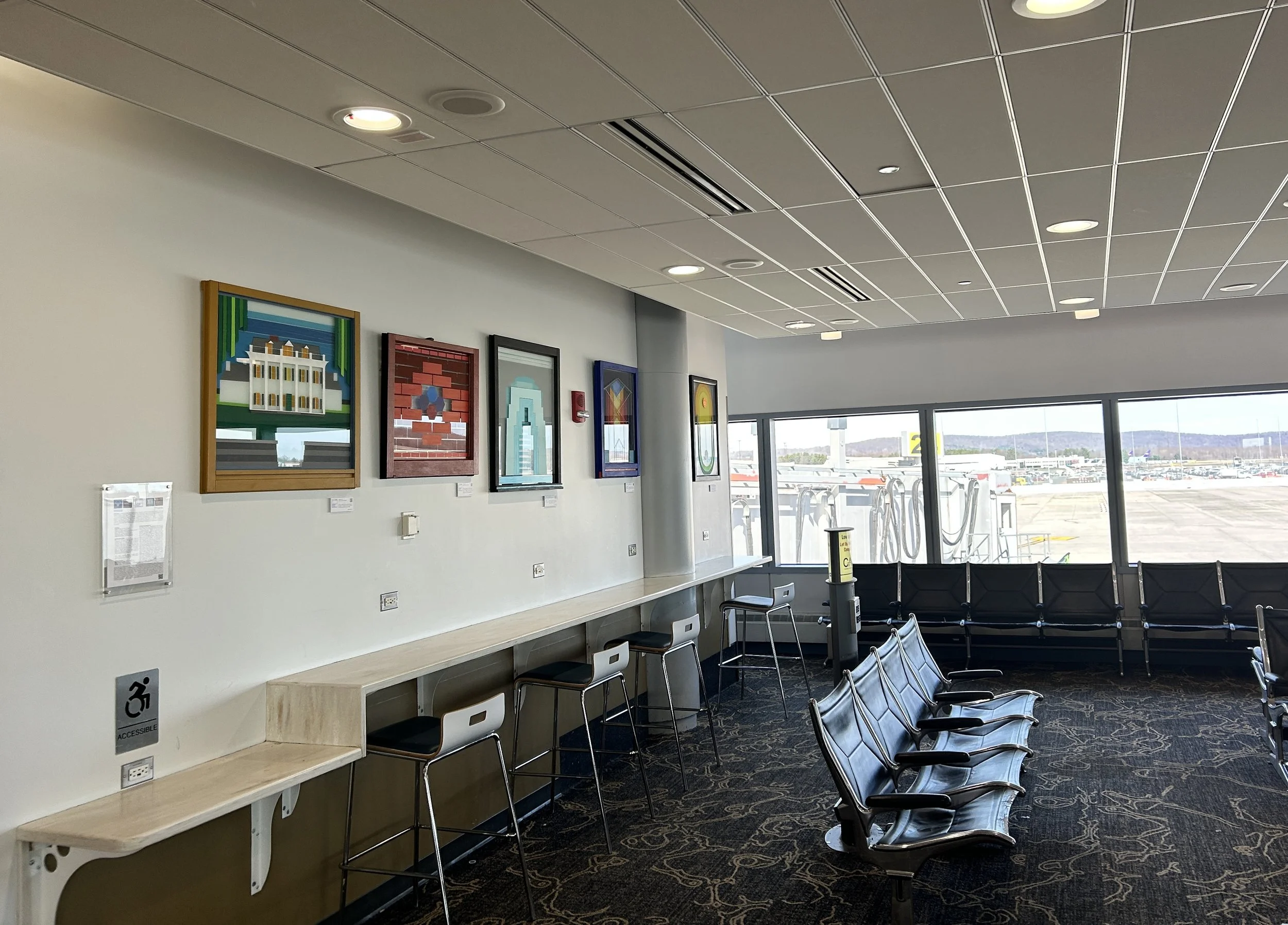 An airport waiting area with black chairs, a row of white chairs along the wall, and artwork on the wall depicting buildings and geometric designs. Large windows show an airport tarmac with airplanes in the background.