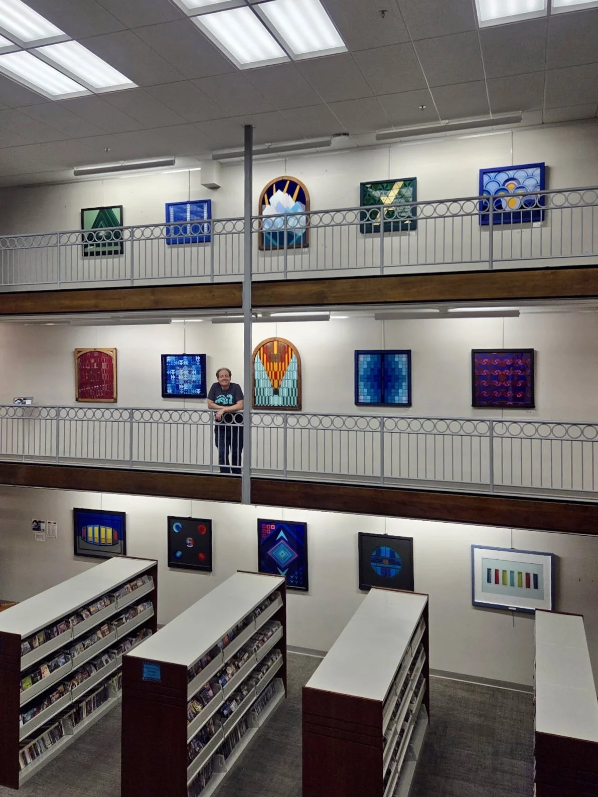 A multi-level art gallery with colorful stained glass artworks on the walls and a man standing on the middle balcony leaning on the railing. The room features bright overhead lighting, and shelves filled with books or DVDs are visible at the bottom.
