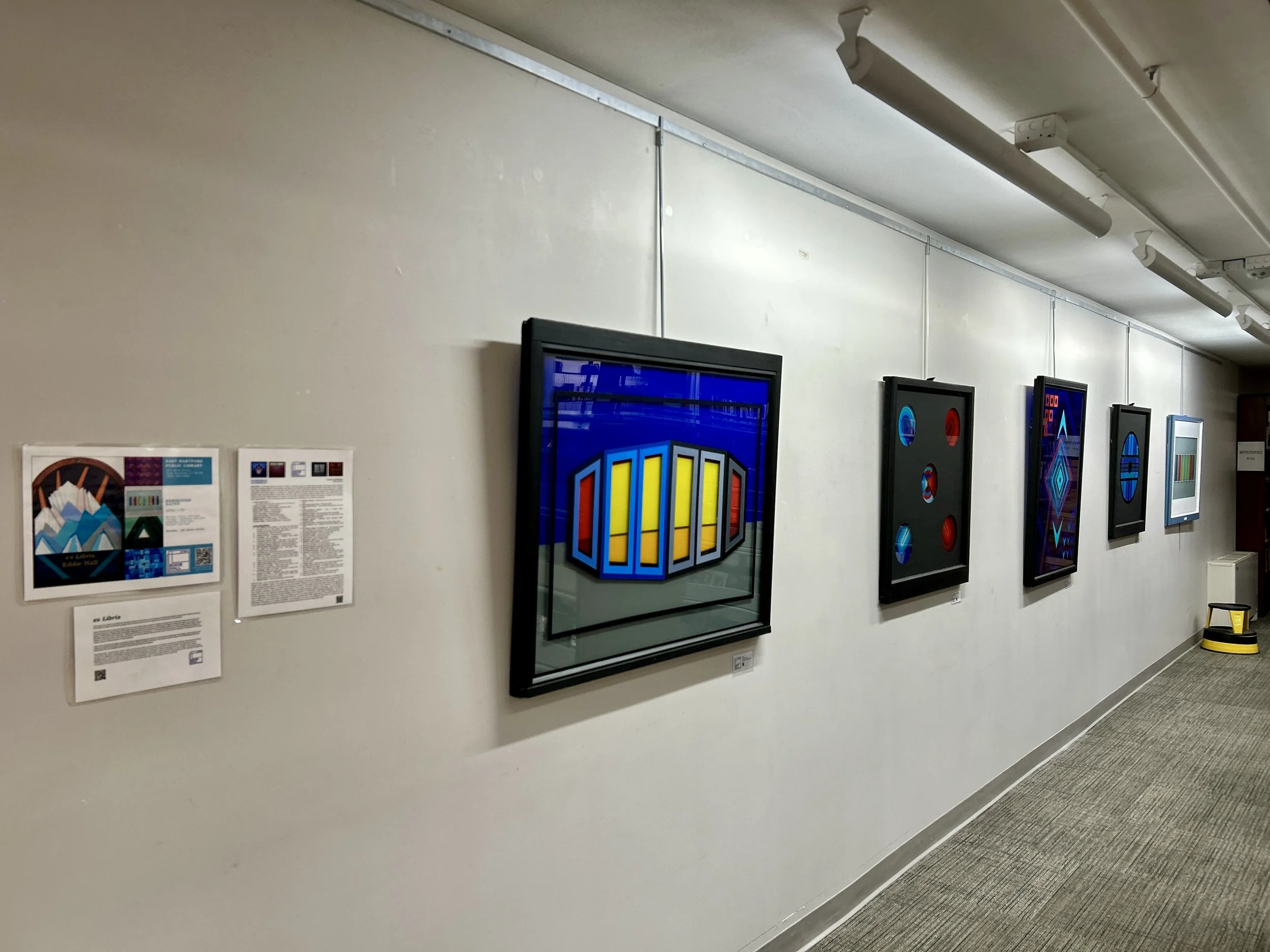 Art gallery wall displaying framed digital artwork and informational posters, with a gray carpeted floor and fluorescent lights overhead.