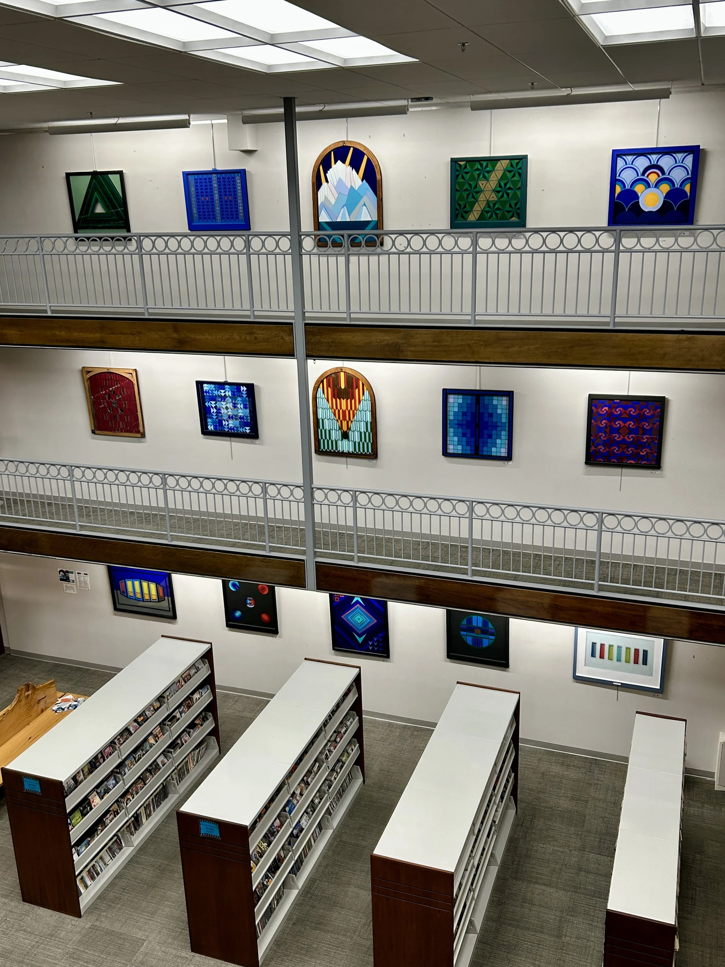 Interior view of a multi-story library with white tables, bookshelves, and artwork on the walls. The upper level has a white railing and colorful abstract paintings, while the lower level has bookshelves with books.