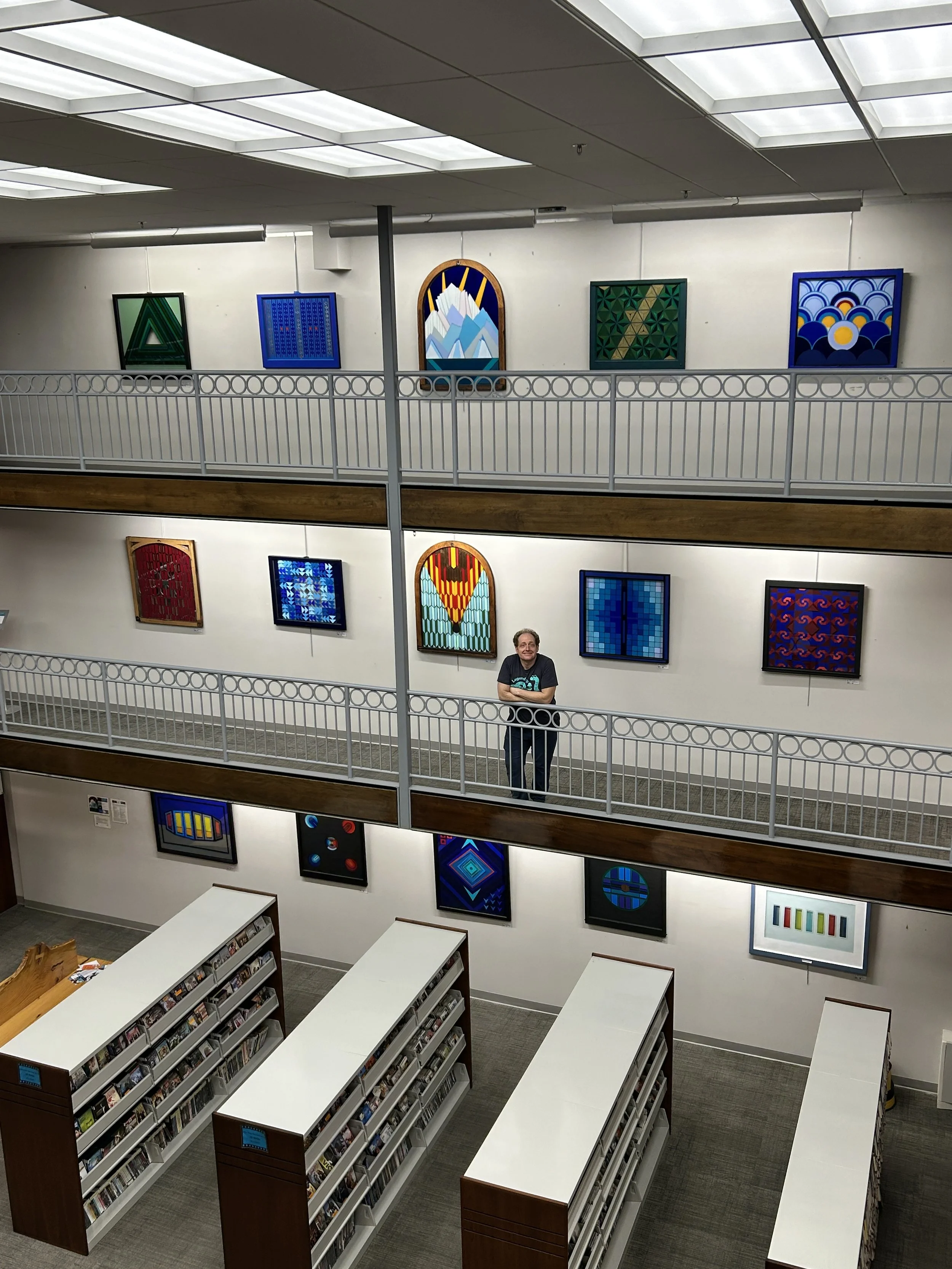 An art gallery with colorful abstract paintings hanging on white walls, a man stands smiling in front of the artwork, and bookshelves are in the foreground.