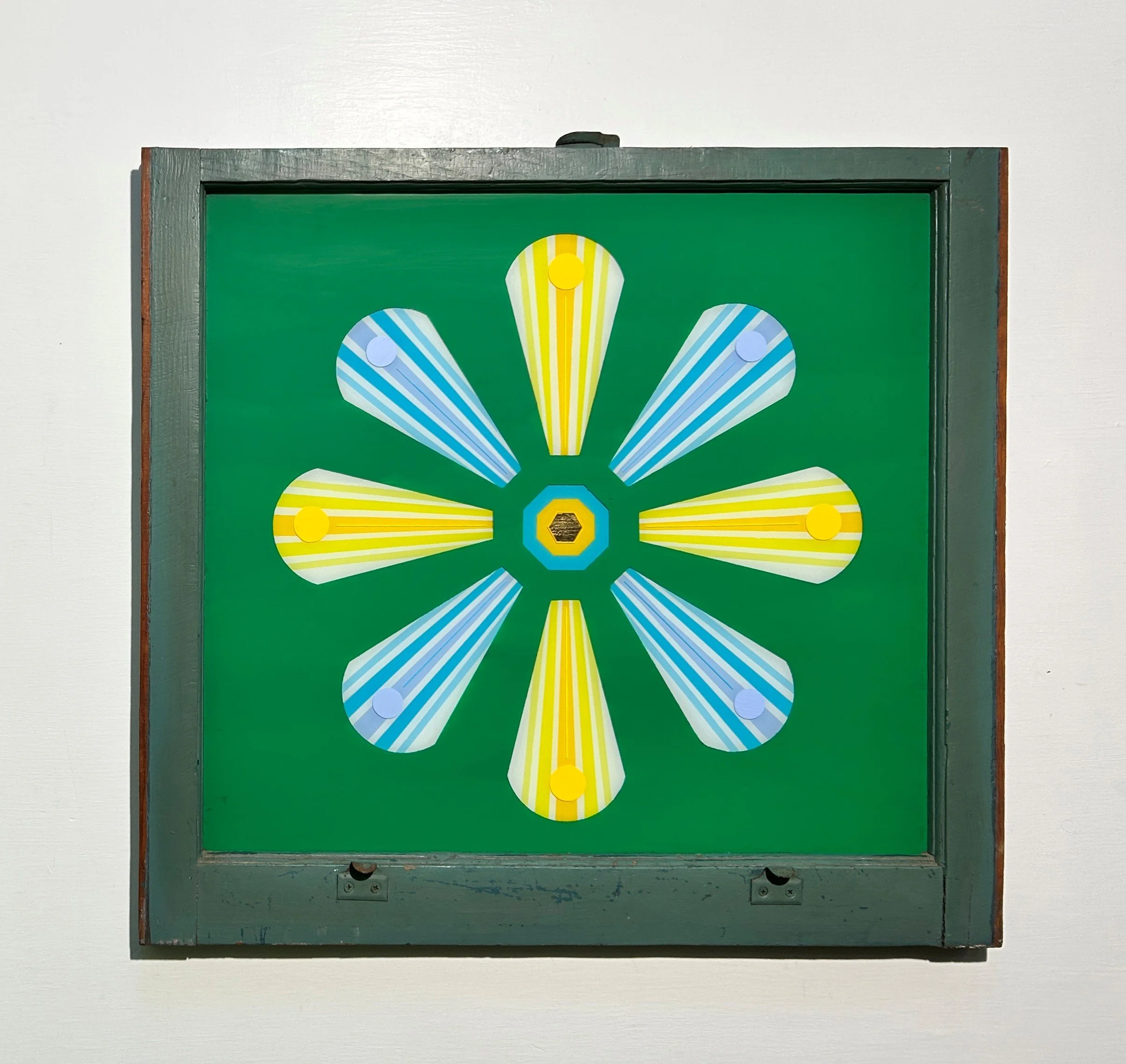 A green board with colorful cutouts in the shape of a flower with blue and yellow petals, pinned to the board, framed with wood, mounted on a white wall.