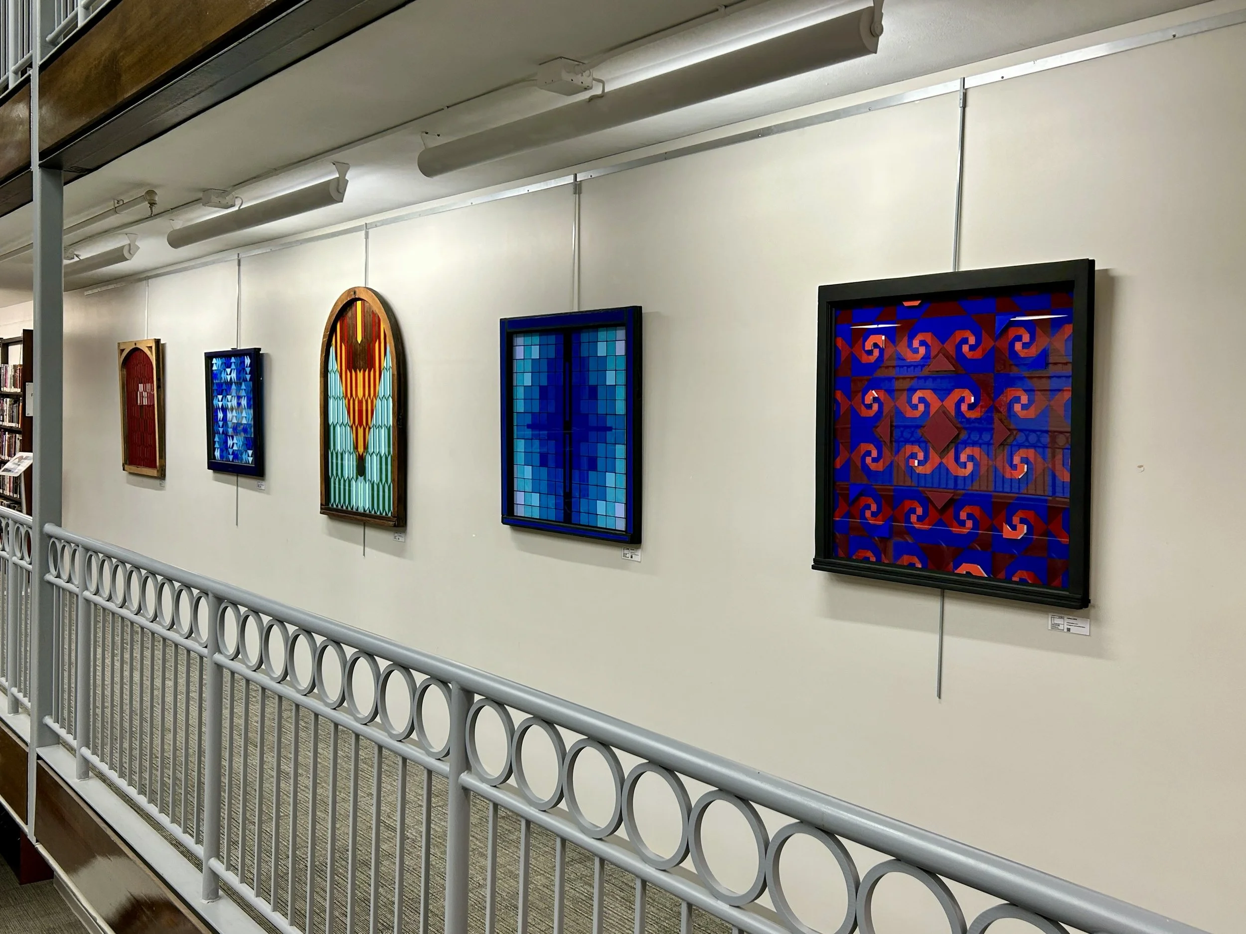 Wall with three framed stained glass art pieces, a bookshelf at the left, and a white railing in the foreground.