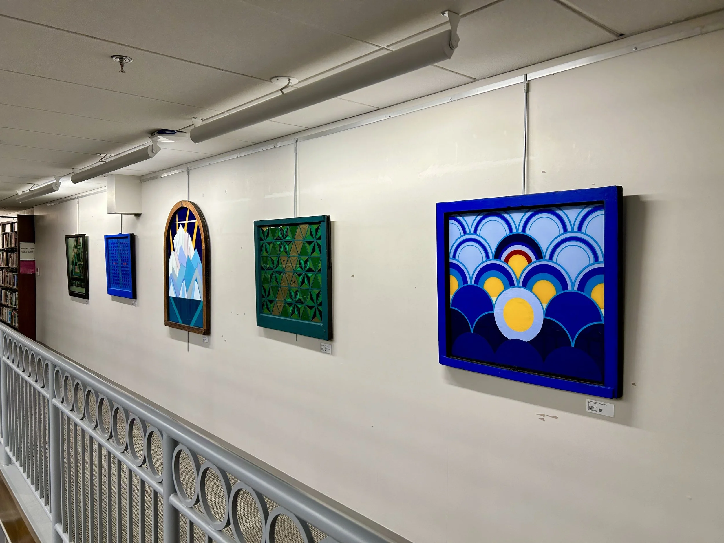 Art gallery wall with four framed geometric and abstract colorful paintings, lit by ceiling track lights in a museum or gallery space.