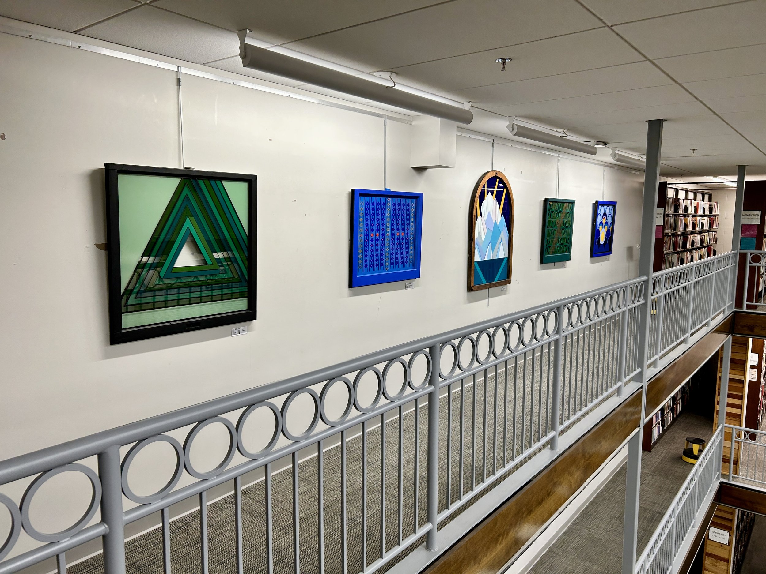 Interior of a library or art gallery with a white wall featuring a series of colorful abstract paintings hung in a row, a white metal railing on a lower level, and bookshelves in the background.