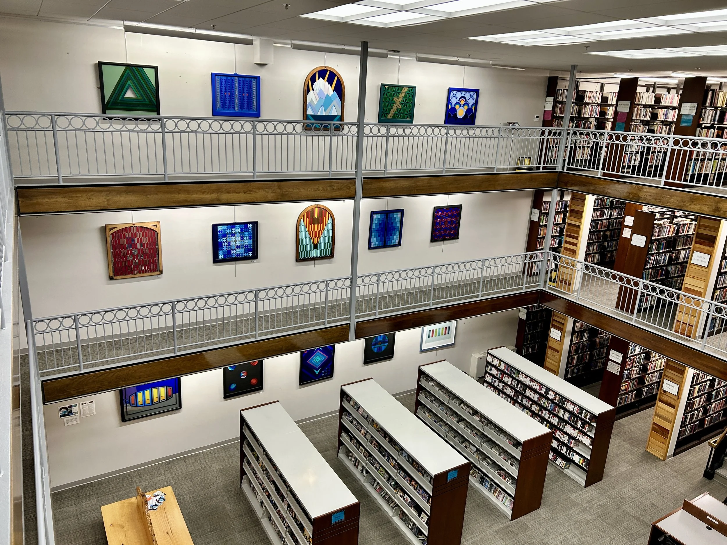 An interior view of a multi-story library with shelves filled with books, decorative paintings hanging on the wall, and white railings along the edges of the open space.