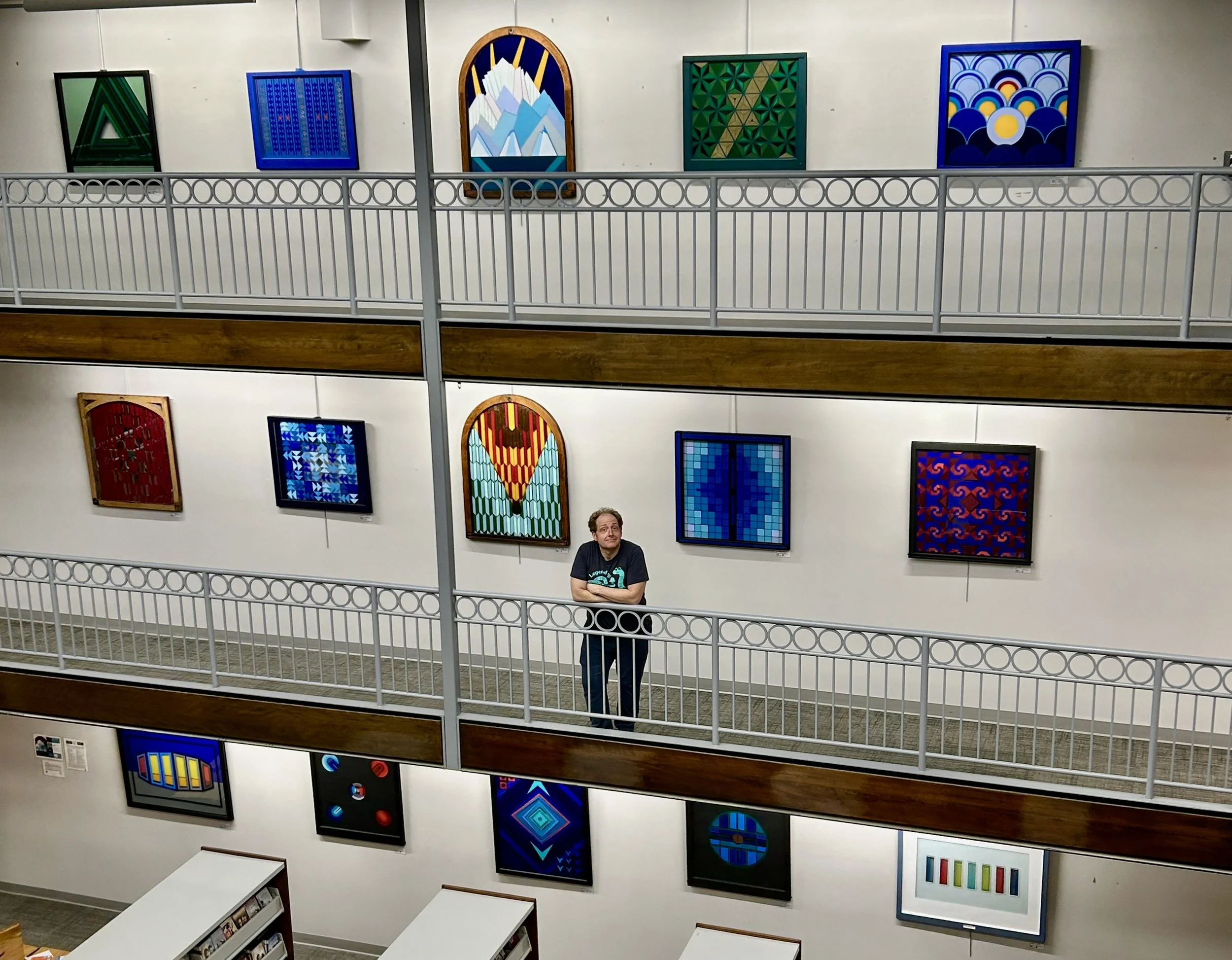 The image shows a man standing on an upper gallery in an art gallery, surrounded by colorful, patterned abstract artworks on the walls. The gallery has a modern design with a metal railing and a wooden accent running horizontally.