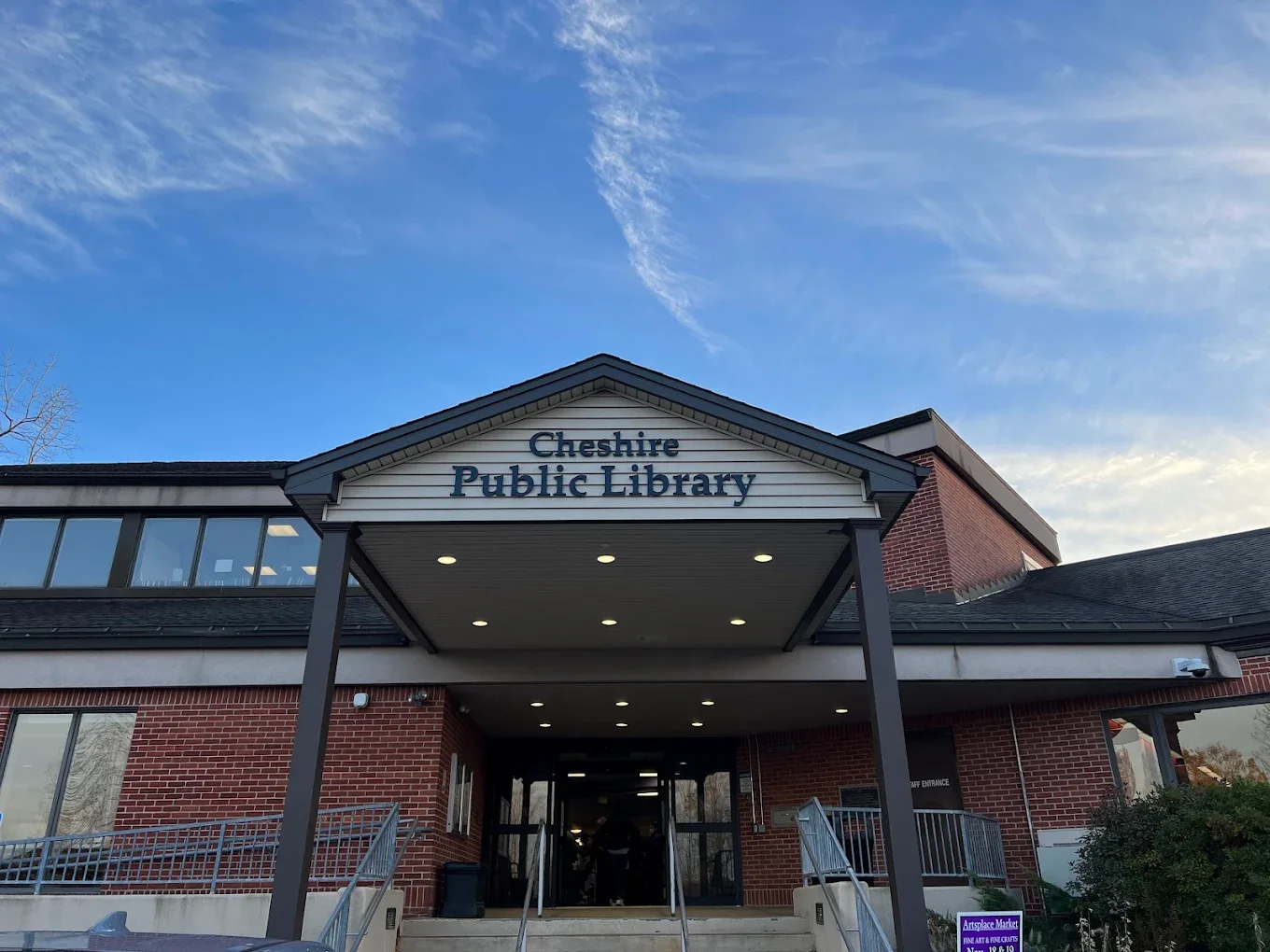 Front view of Cheshire Public Library building with brick exterior, blue sky, and overhead lights at the entrance.