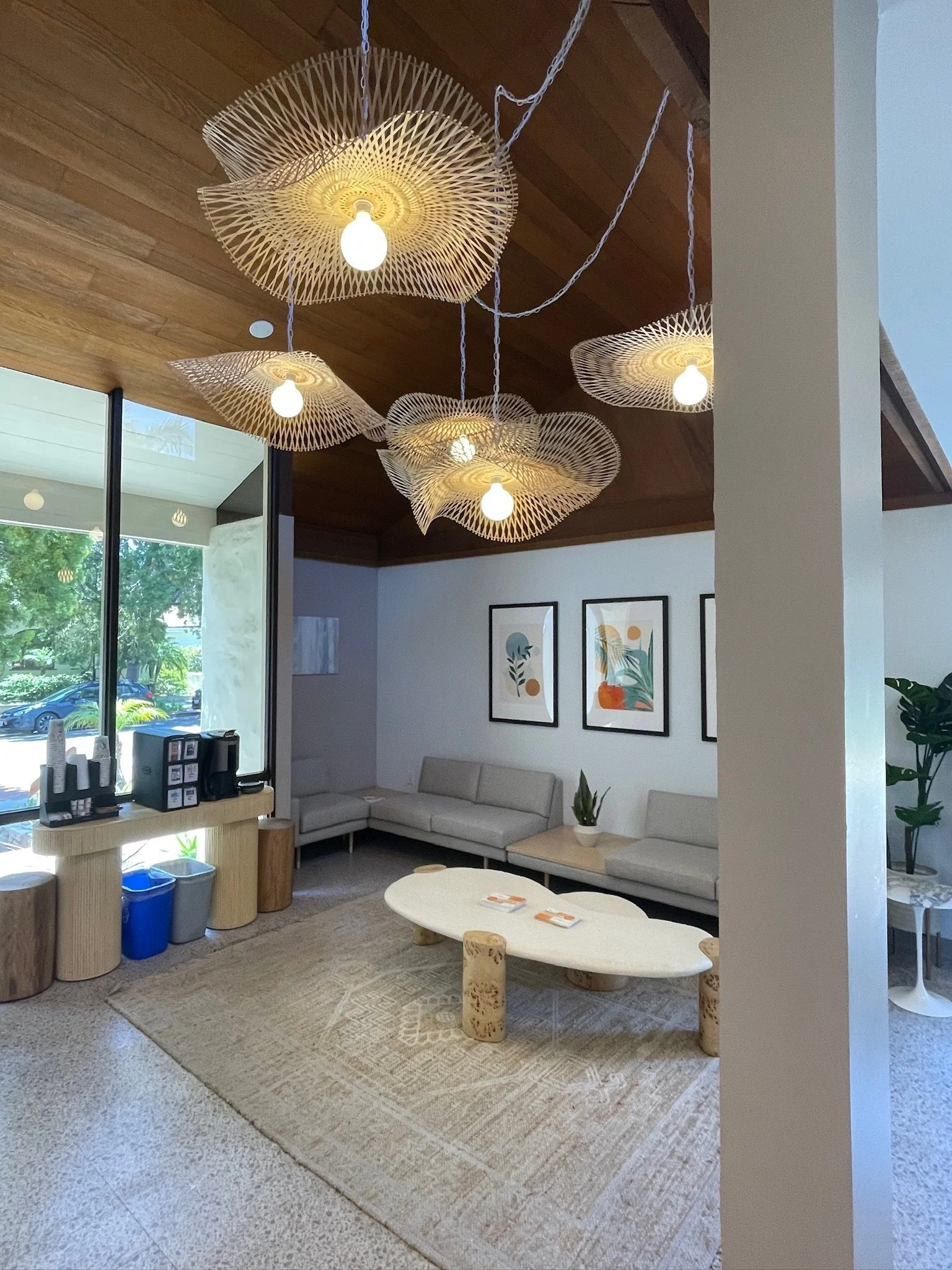 Modern hotel lobby with beige sectional sofa, white oval coffee table, decorative paintings on the wall, wooden ceiling with hanging wicker light fixtures, and large windows showing trees outside.