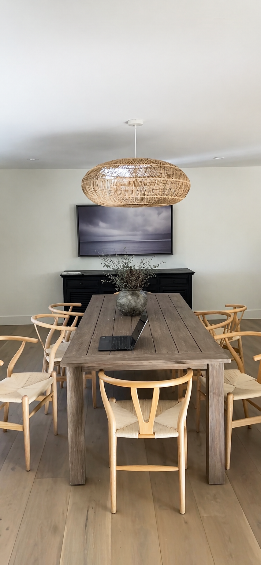 A wooden dining table with a laptop on it, surrounded by eight light-colored wooden chairs with woven seats, a black sideboard, a large framed landscape photo of a cloudy sky over water on the wall, a vase with greenery, and a woven pendant light han
