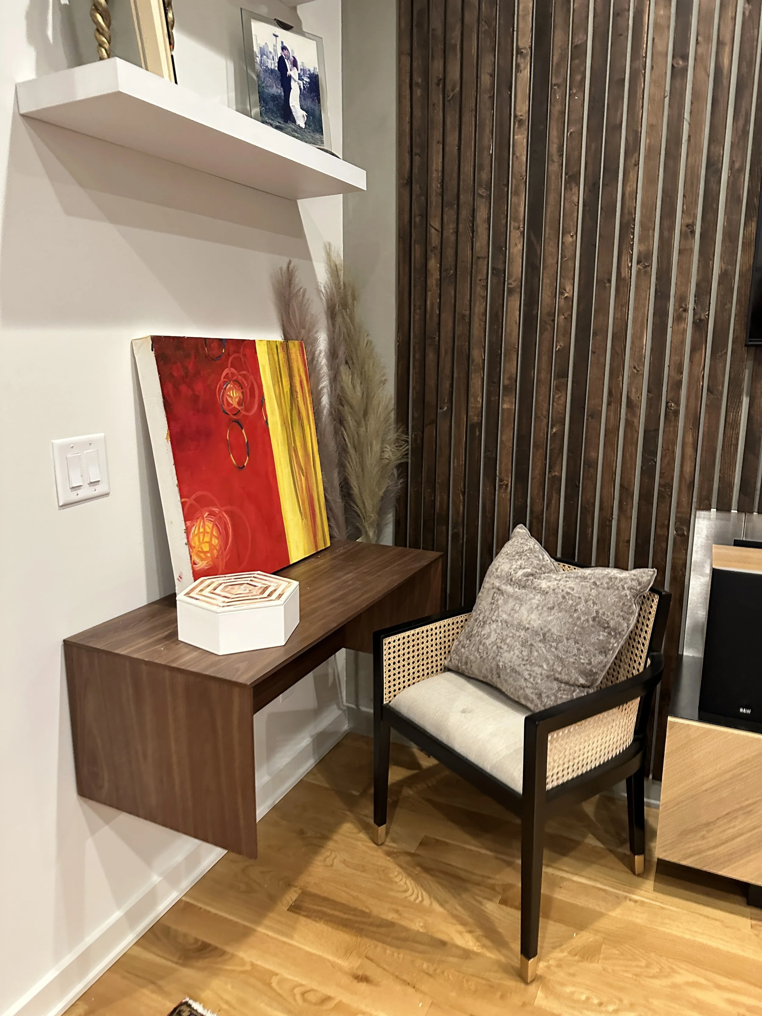 A corner of a room with a wooden floor and a white wall. A wooden desk with a decorative box and colorful abstract painting is next to a chair with black frame and beige cushions, with a light gray pillow. In the background, there is a wooden slat wa