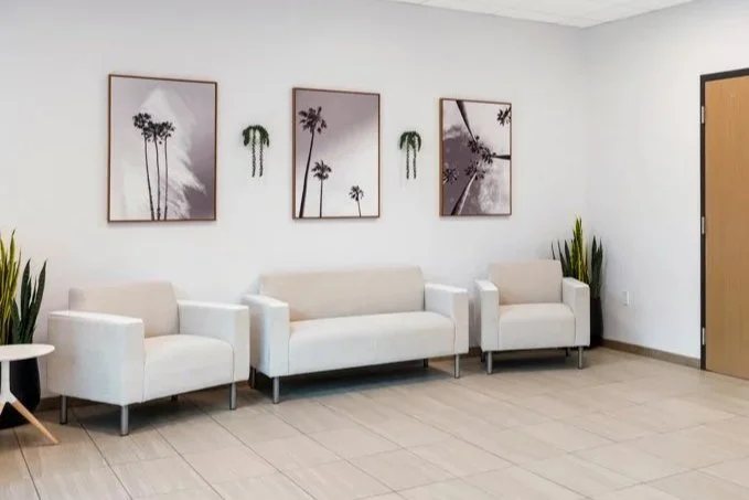 A waiting room with three white sofas, a small side table, potted plants, and framed palm tree artwork on the wall.