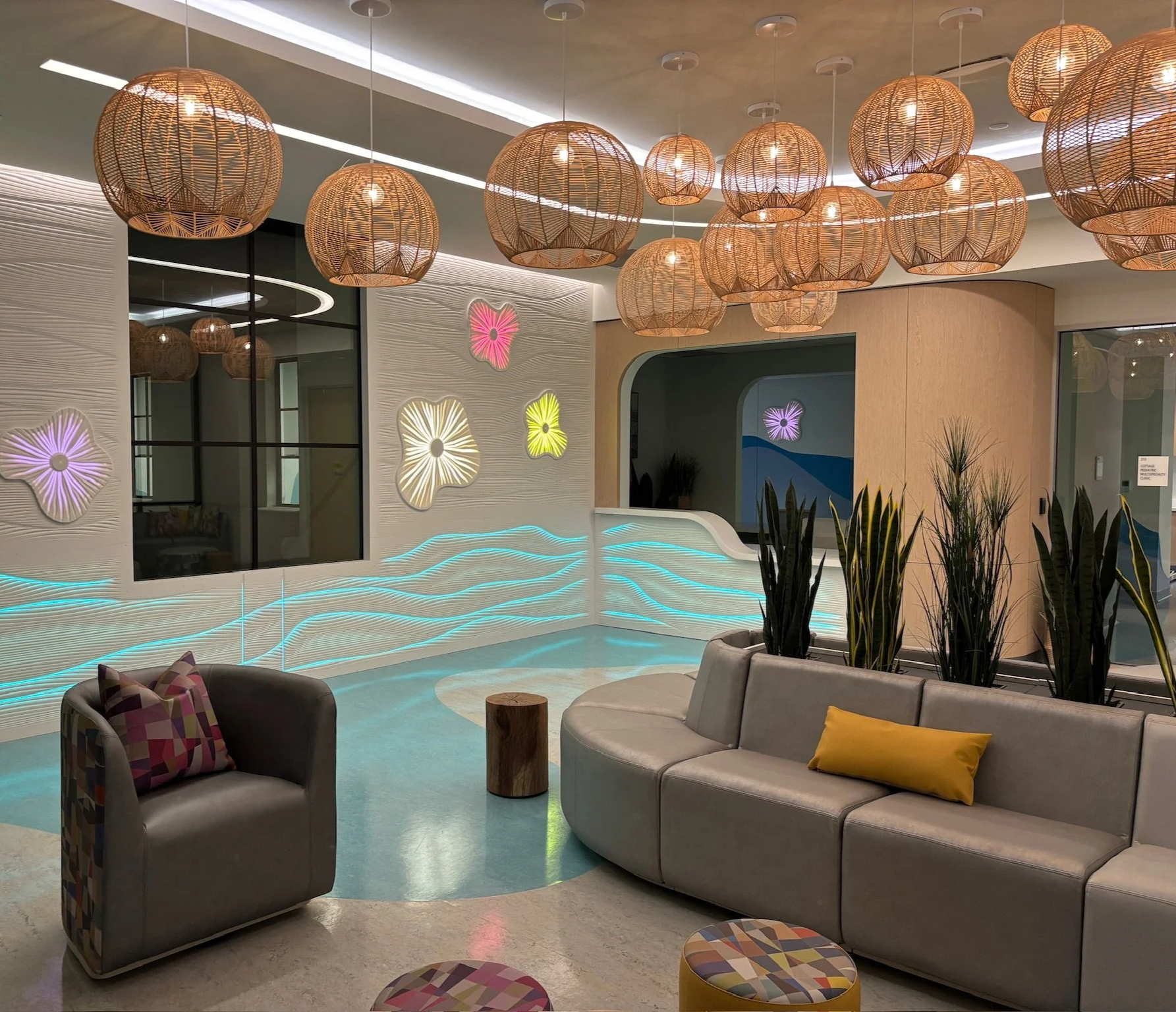 Modern lounge area with hanging wicker light fixtures, colorful illuminated floral wall decorations, a curved beige sofa with pillows, a black armchair with a patterned pillow, and several round stools, some with multicolored geometric patterns. Ther